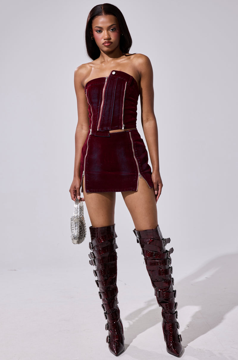 A woman models the ASYMMETRIC ZIP CORSET— a strapless dark red velvet mini dress with front zippers—paired with matching knee-high boots with buckles. She holds a silver handbag and stands against a plain light gray background. 4