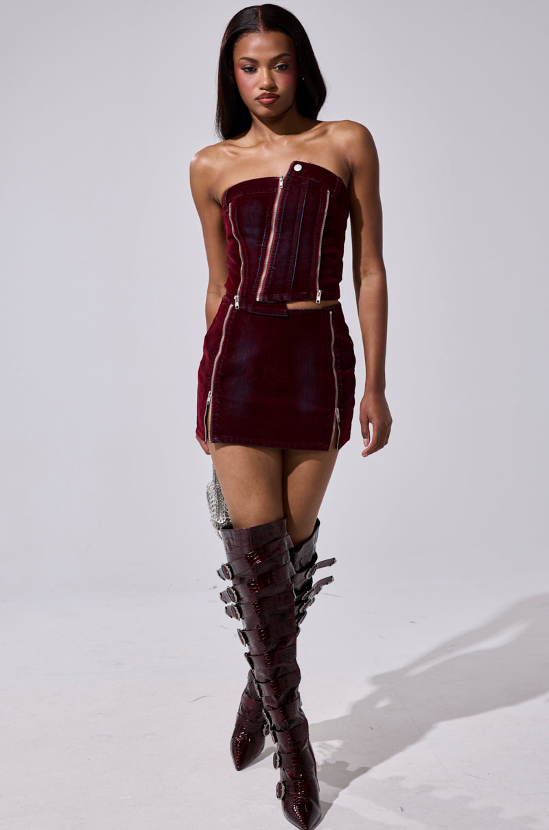 A woman with straight dark hair confidently walks against a plain light background, wearing the ASYMMETRIC ZIP CORSET—a strapless burgundy denim mini dress with zipper details and paired with matching high-heeled, multi-buckle boots. 5
