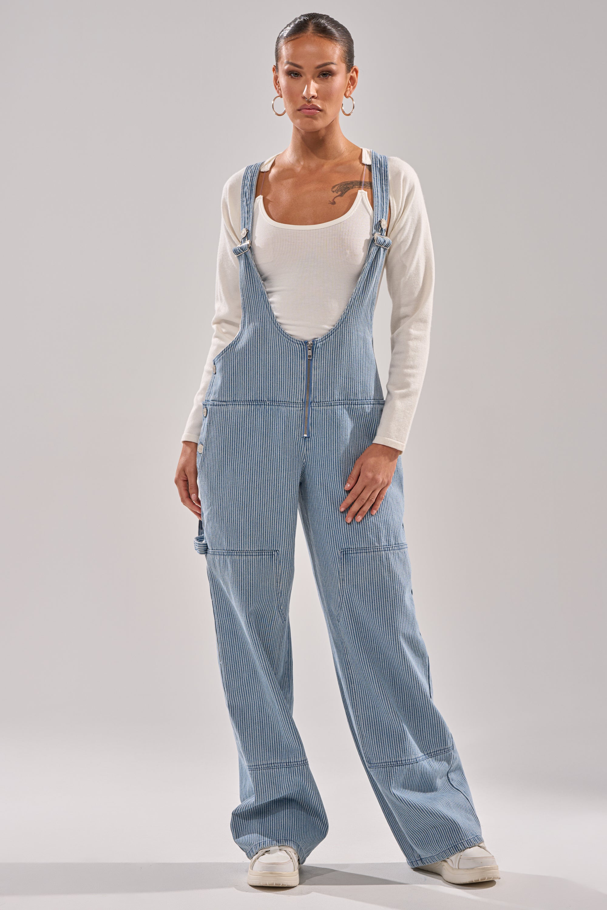 A person faces forward in HAMPTONS STRIPED CARPENTER OVERALLS over a fitted white long-sleeve shirt and white sneakers, hair pulled back with large hoop earrings—a modern take on women’s workwear. 0