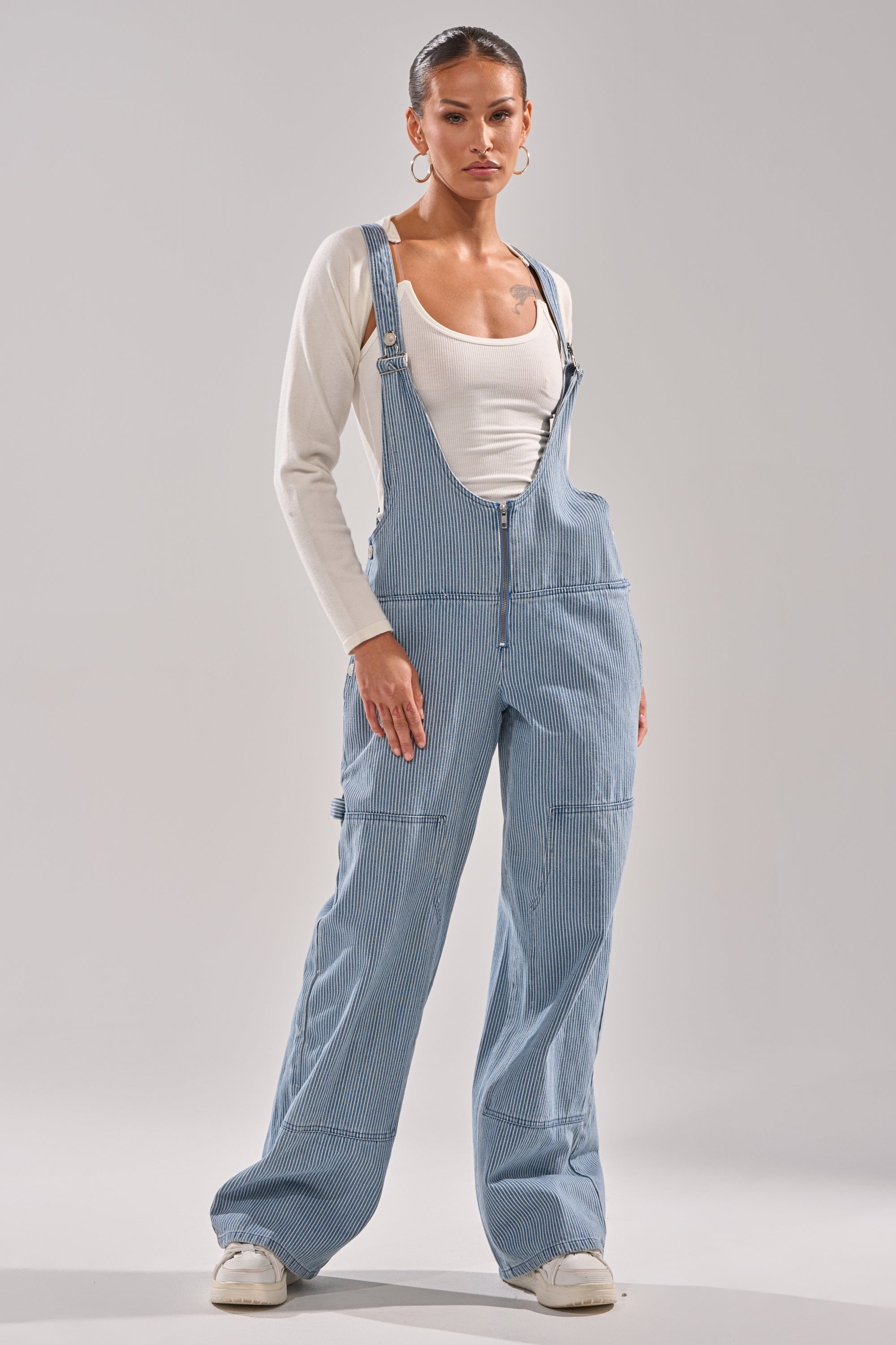A woman stands confidently in HAMPTONS STRIPED CARPENTER OVERALLS, layered over a fitted white long-sleeve shirt and paired with white sneakers, against a plain light gray background. 1
