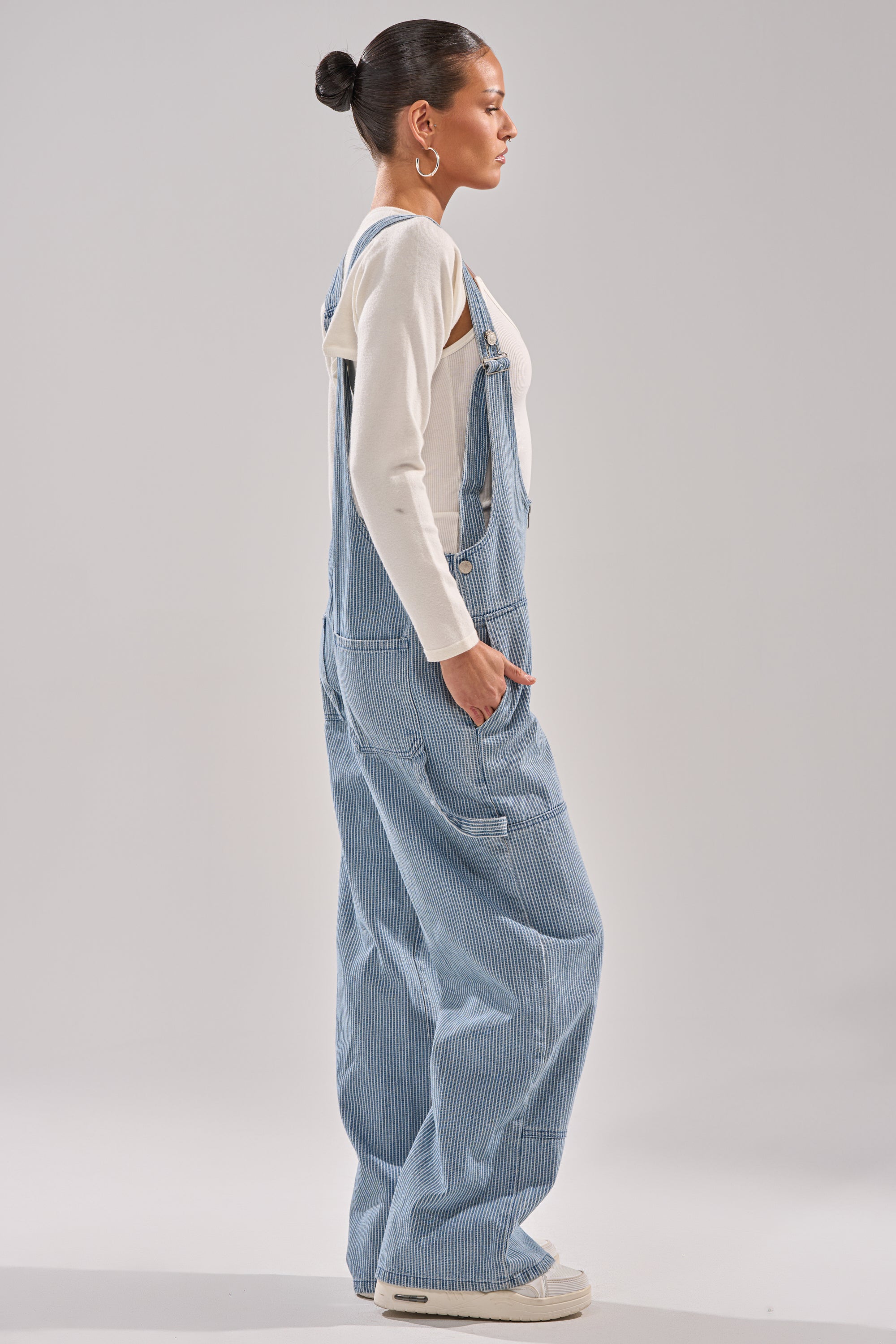 A person stands in profile wearing HAMPTONS STRIPED CARPENTER OVERALLS over a cream long-sleeve shirt, with hands in pockets, hair in a bun, and white sneakers against a plain light background. 2