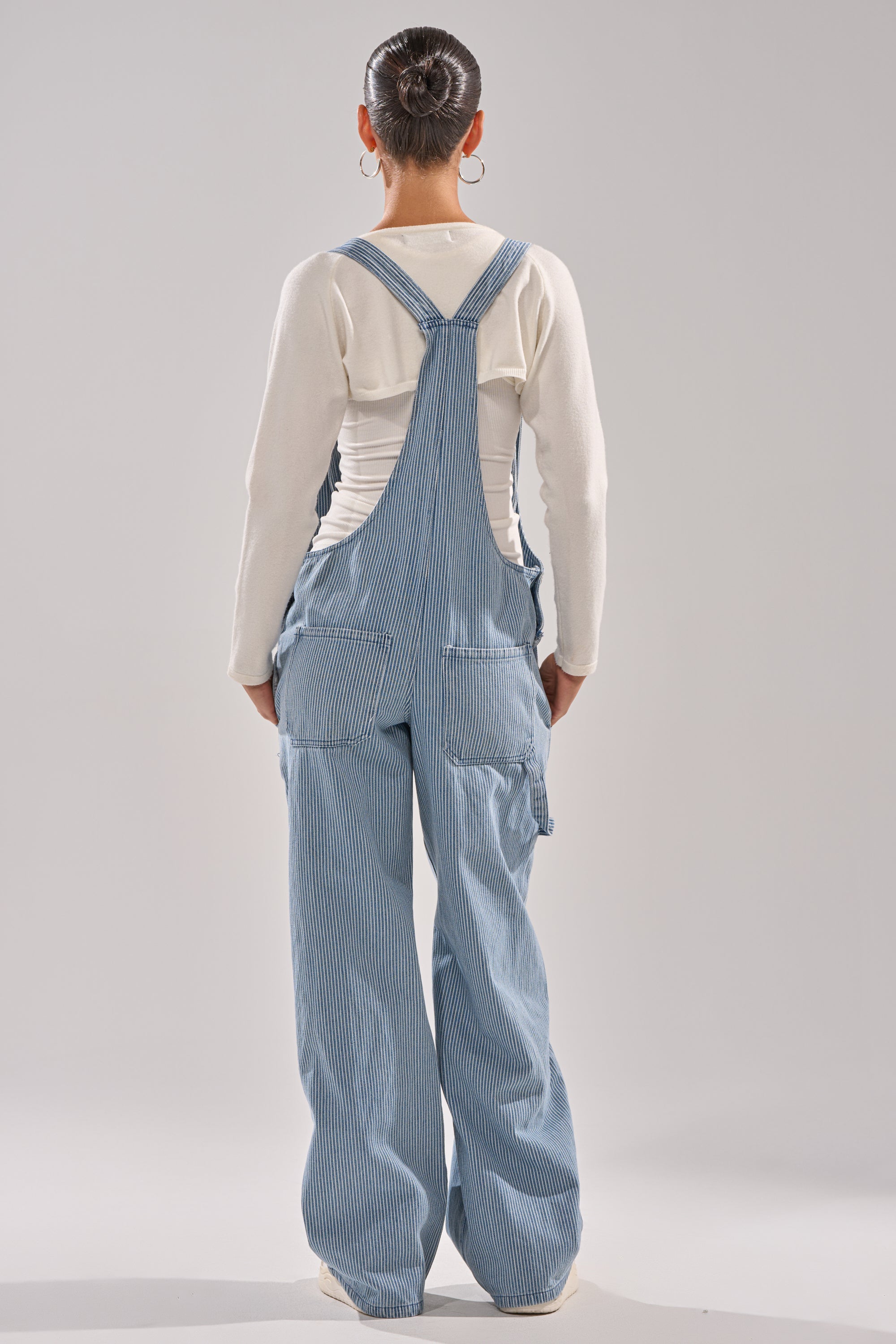 A person with their hair in a neat bun stands facing away against a light gray background, wearing a white long-sleeve shirt paired with HAMPTONS STRIPED CARPENTER OVERALLS featuring wide legs. 3