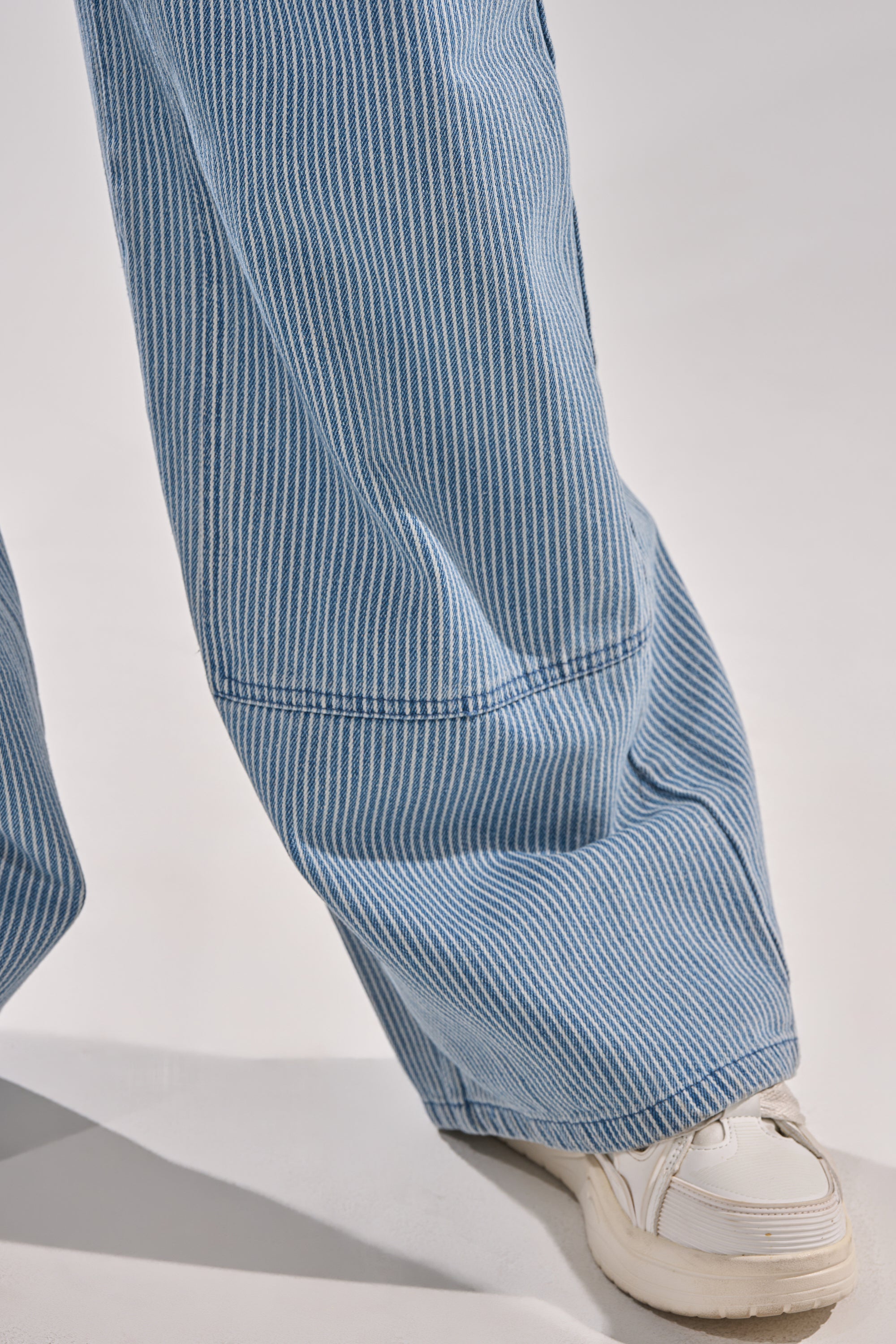 Close-up of a person wearing HAMPTONS STRIPED CARPENTER OVERALLS in blue and white stripes with wide legs, paired with white sneakers, standing on a light surface. 5