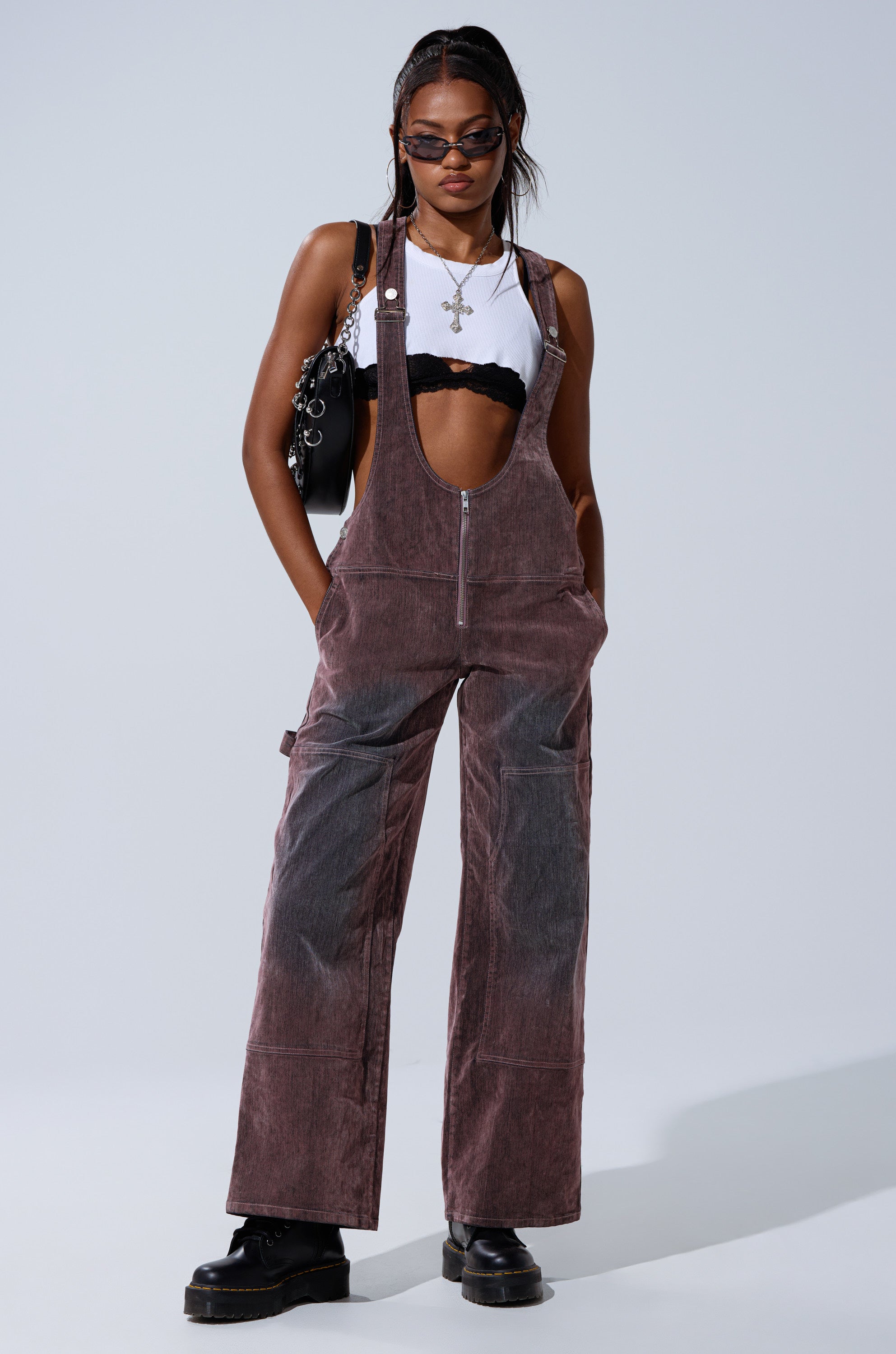 A woman poses confidently in DOUBLE KNEE TEXTURED DENIM OVERALLS over a white crop top and black bralette, styled with sunglasses, layered necklaces, a black handbag, and chunky black shoes against a plain background.