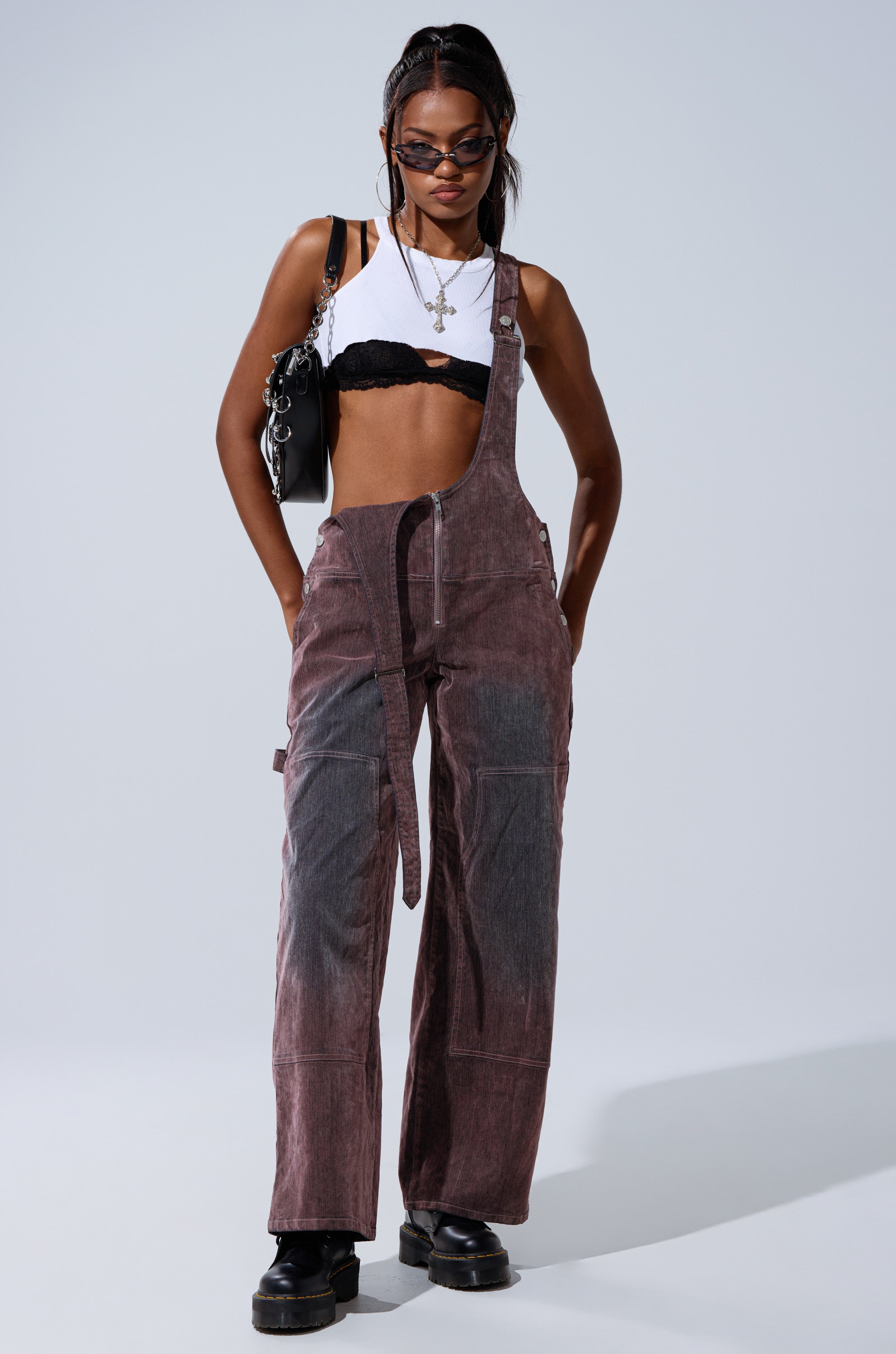 A woman in DOUBLE KNEE TEXTURED DENIM OVERALLS with one strap undone, sunglasses, a white crop top over a black bralette, chunky black shoes, and a black purse stands hands-in-pockets against a plain background.
