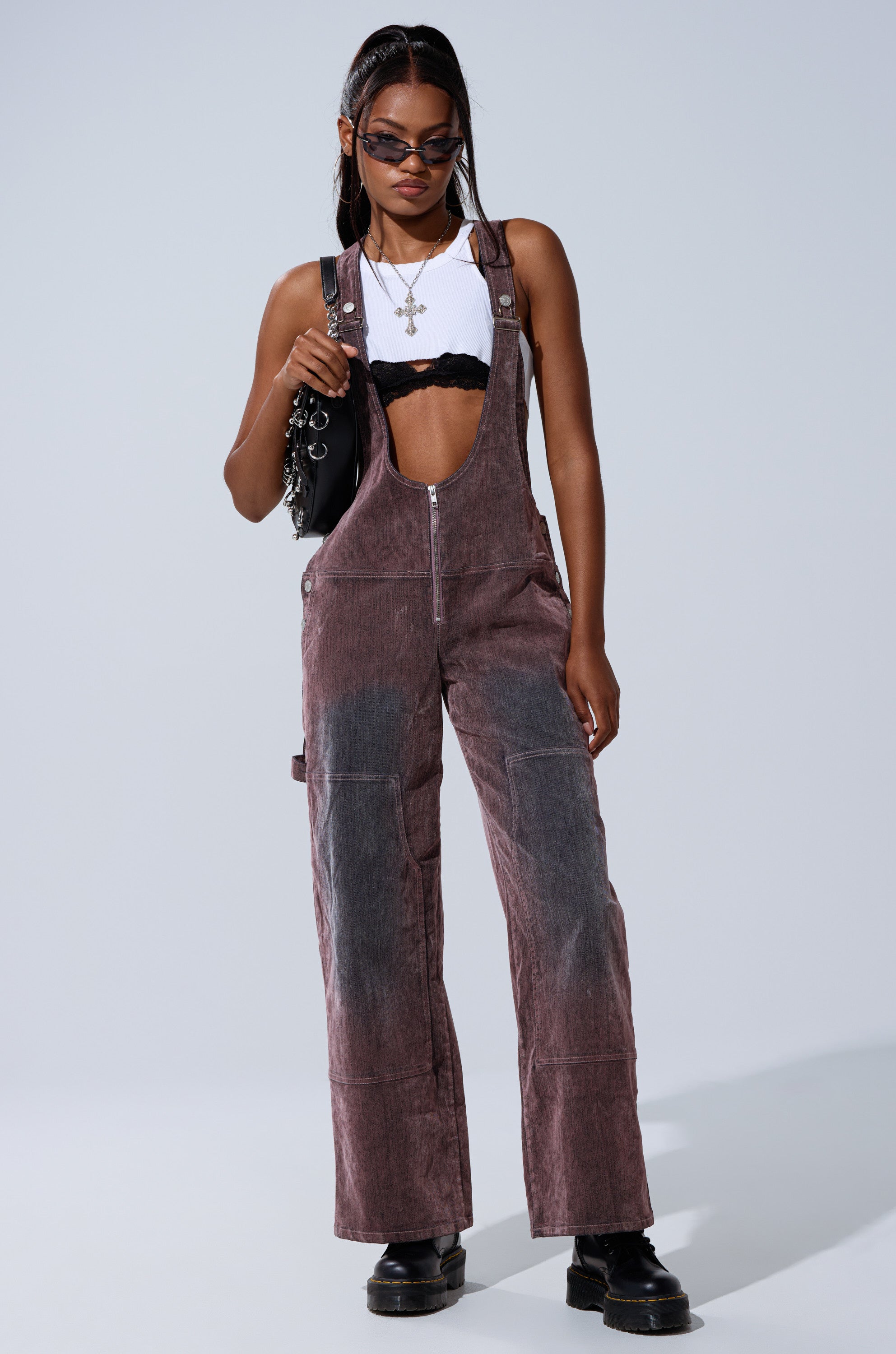 A woman stands confidently against a plain background wearing DOUBLE KNEE TEXTURED DENIM OVERALLS over a white crop top and black bralette, styled with sunglasses, layered necklaces, a black bag, and chunky black shoes.