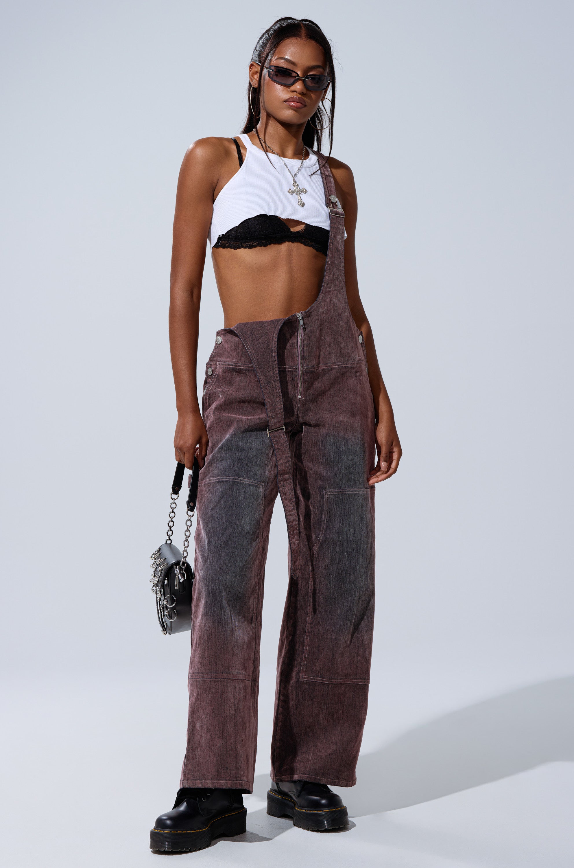 A woman stands confidently against a plain background, wearing sunglasses, a white crop top over a black bra, DOUBLE KNEE TEXTURED DENIM OVERALLS with one strap, black platform shoes, and holding a small handbag.