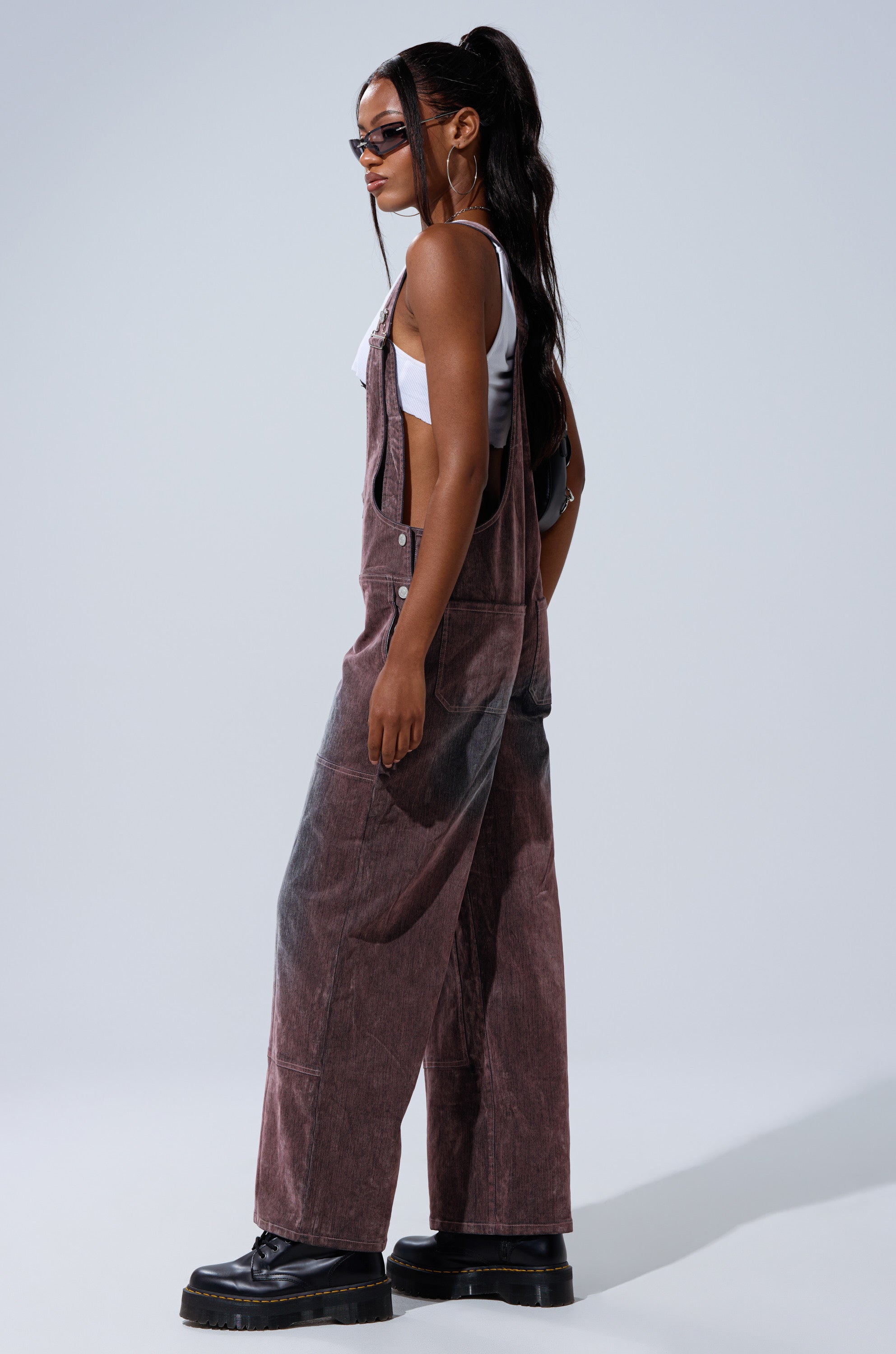 A woman wearing DOUBLE KNEE TEXTURED DENIM OVERALLS, a white crop top, black platform shoes, and sunglasses stands sideways against a plain light gray background.