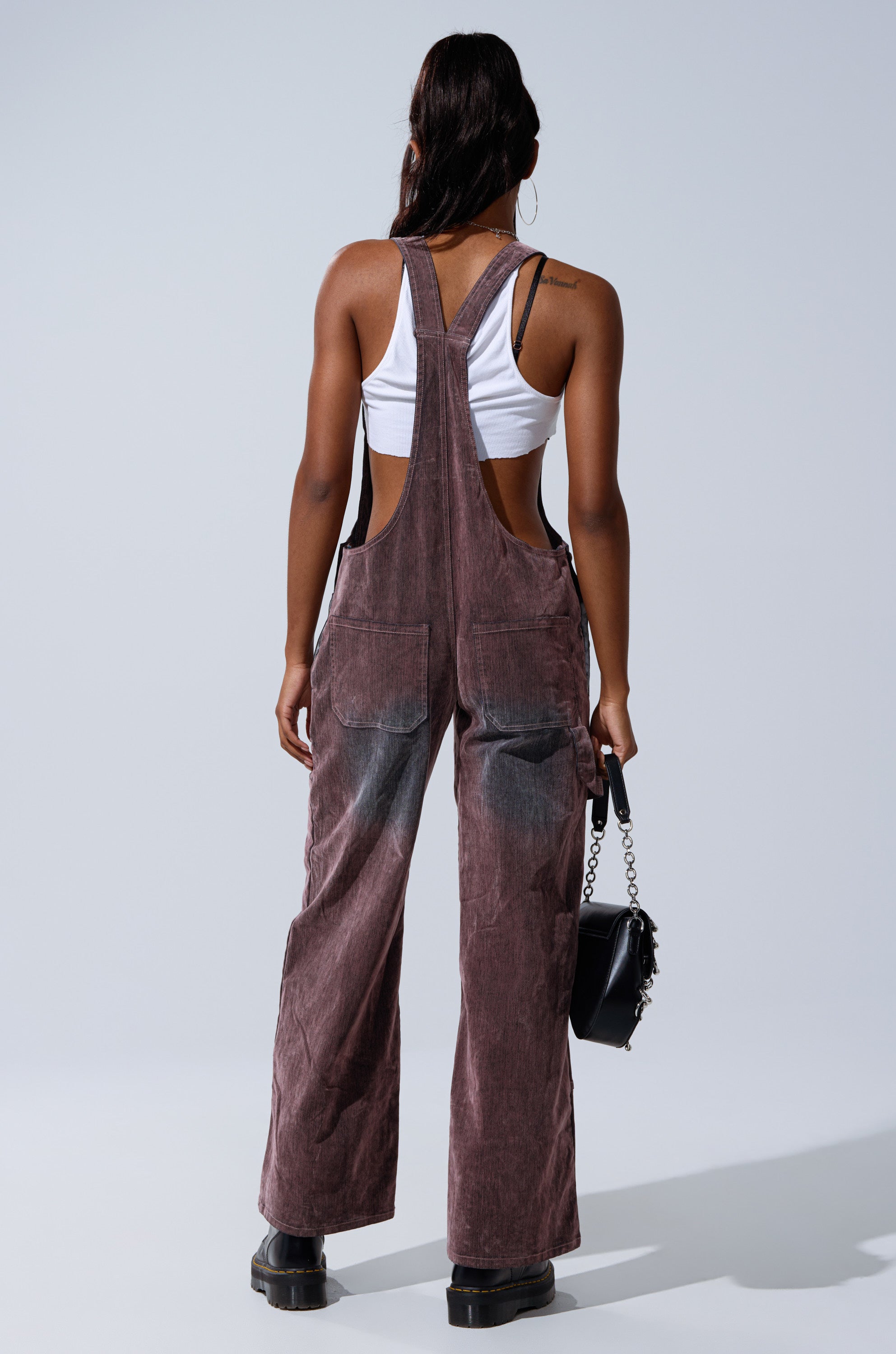 A person stands facing away, wearing a white crop top and DOUBLE KNEE TEXTURED DENIM OVERALLS with wide legs and large back pockets, paired with black boots and a chain-strap black handbag.