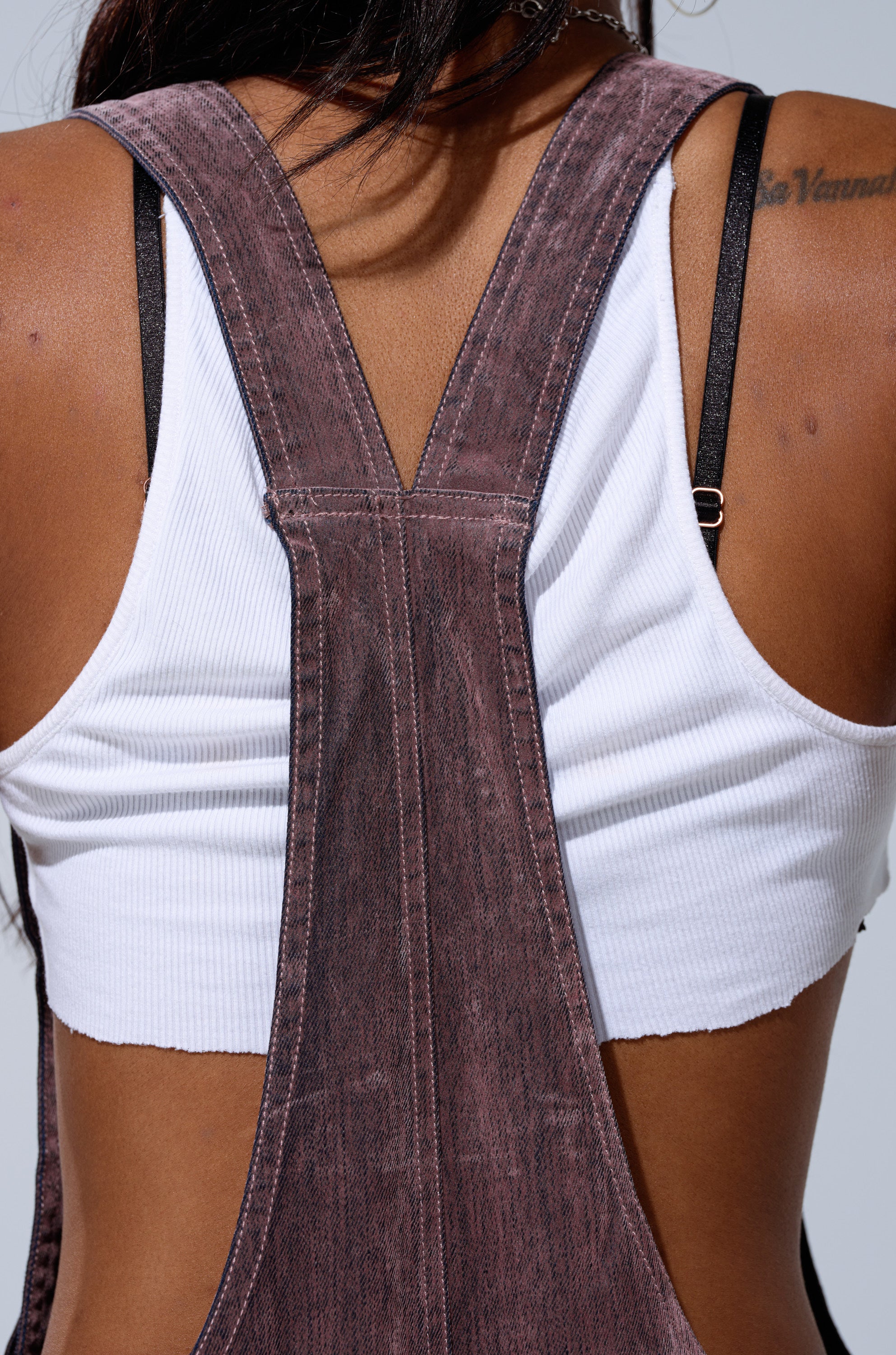 A close-up shows a person’s back in a white ribbed crop top and DOUBLE KNEE TEXTURED DENIM OVERALLS. Medium brown skin, visible shoulder straps, and a tattoo on the right shoulder accent this women’s casual denim style.