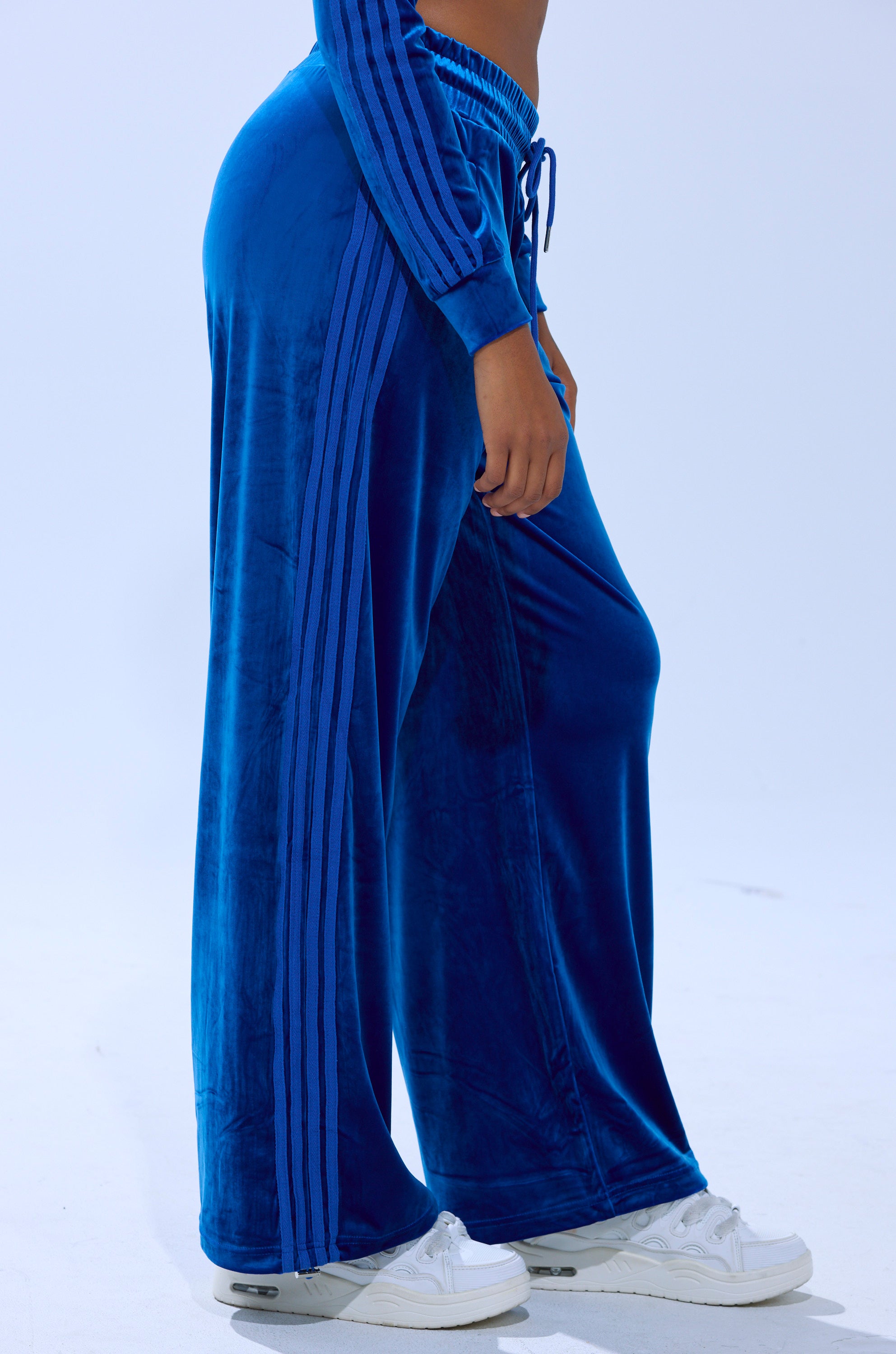 A person models the ELECTRIC BLUES WIDE LEG JOGGER—high-rise blue velour pants with side stripes and a matching jacket—styled with white sneakers against a plain light background.