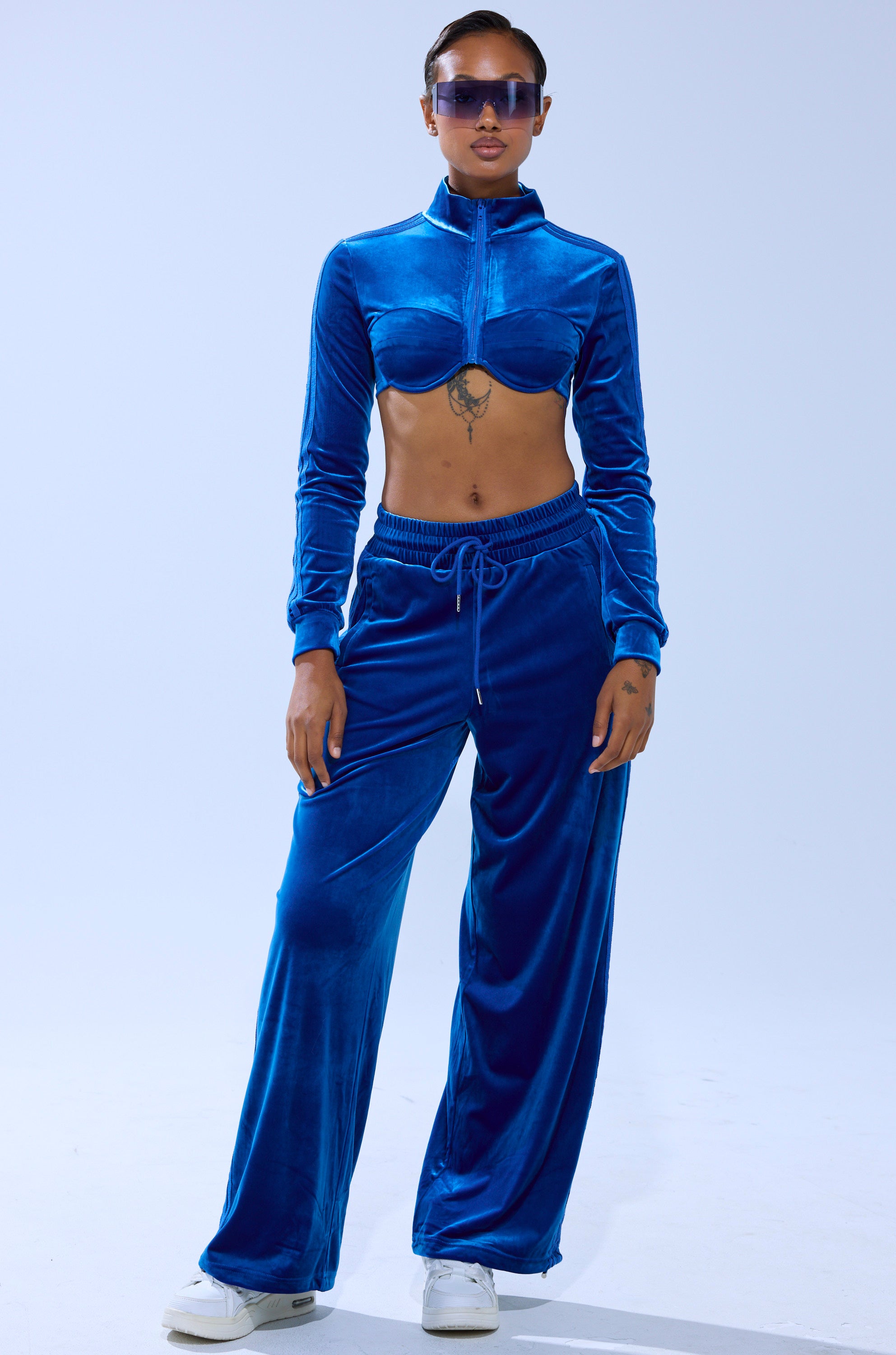 Wearing the ELECTRIC BLUES WIDE LEG JOGGER and a sapphire-blue cropped zip-up top, a tattooed person in white sneakers and sunglasses stands confidently against a plain light background.