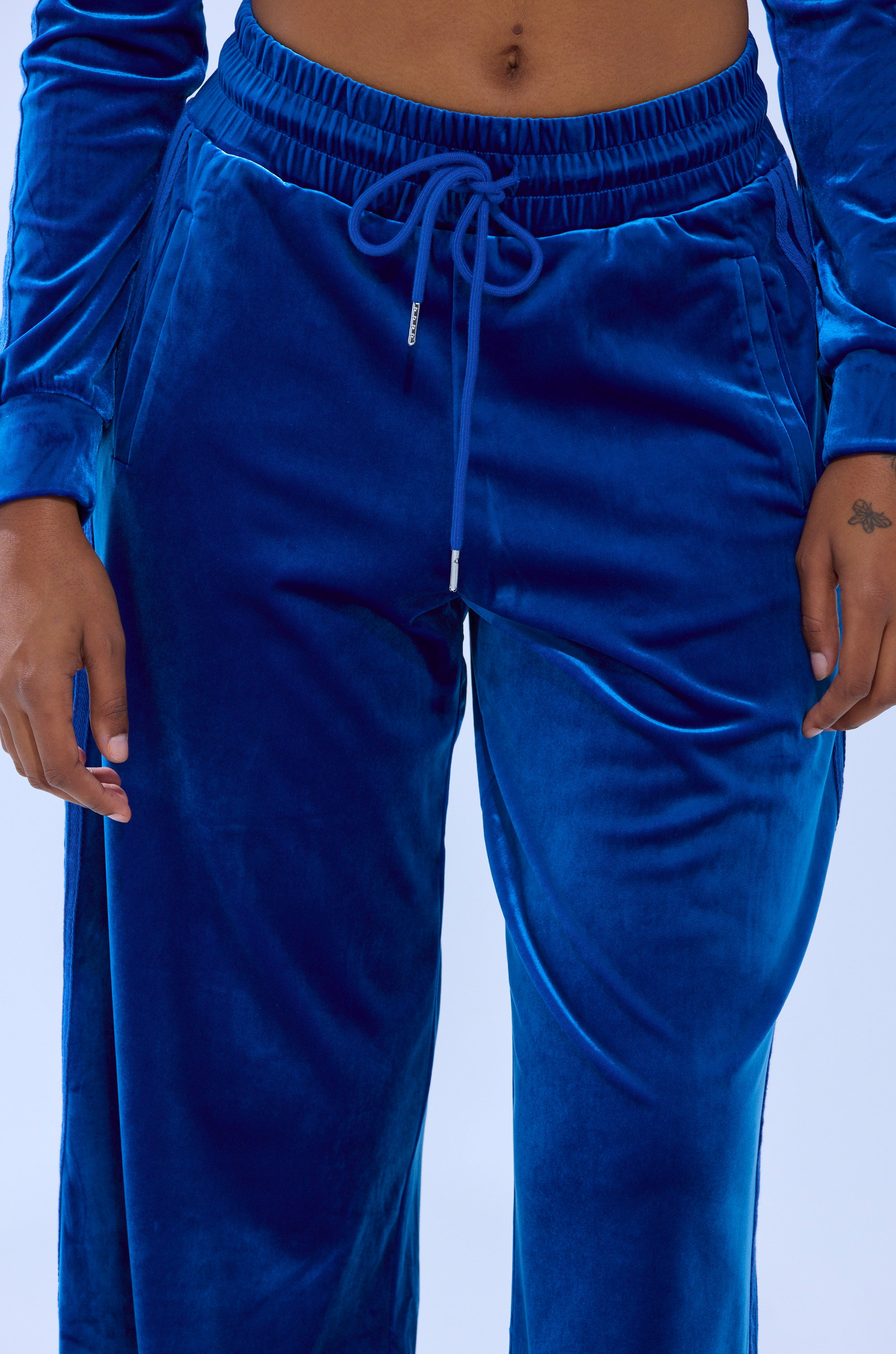 A person wears the ELECTRIC BLUES WIDE LEG JOGGER in sapphire-blue velour, paired with a matching long-sleeve top. Only their torso and upper legs are shown, with hands resting by their sides.