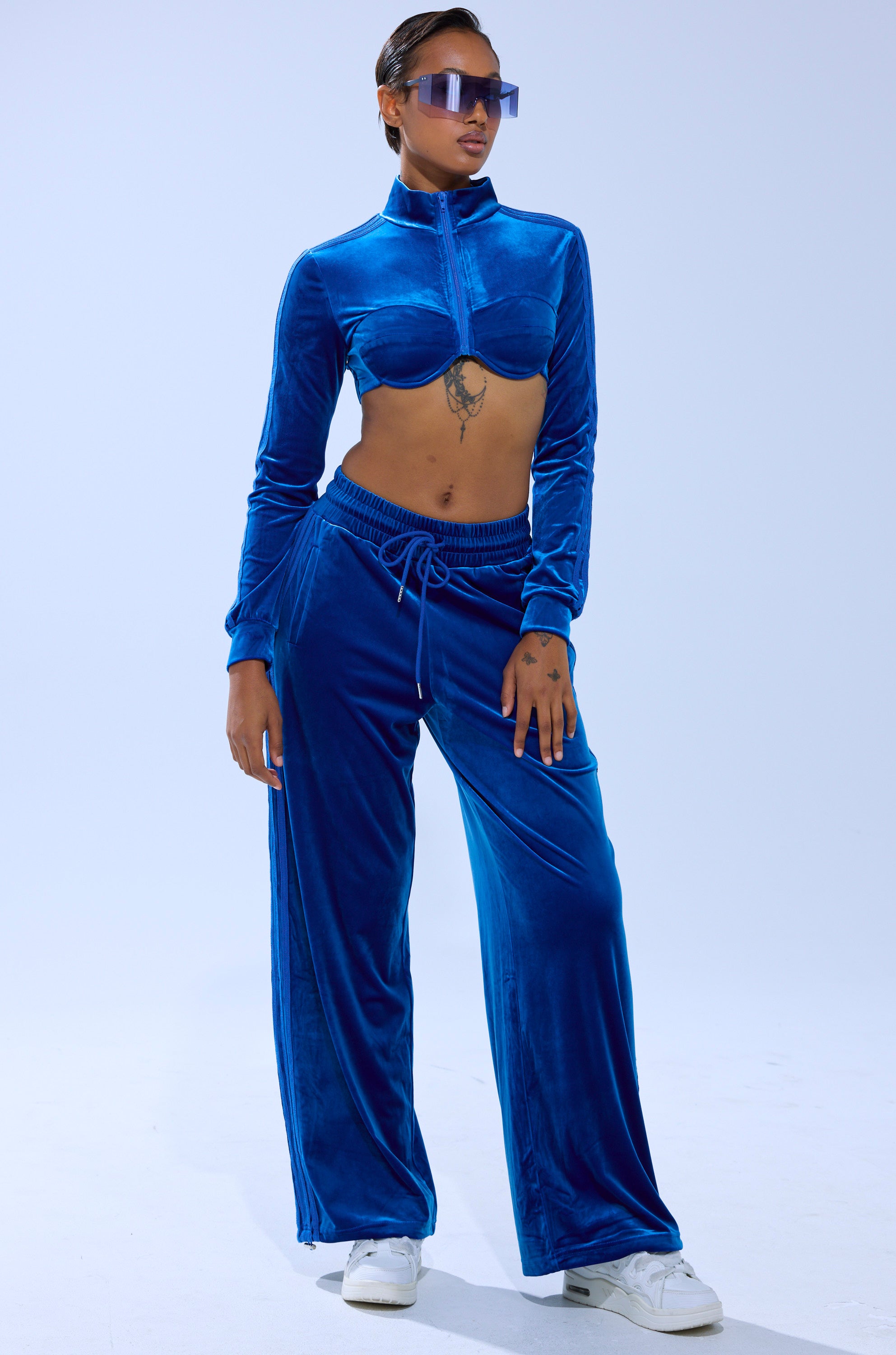A person in the ELECTRIC BLUES CROP ZIP UP SWEATSHIRT, a sapphire-blue velour cropped zip-up top with a bustier-inspired design and wide-leg pants, paired with white sneakers and sunglasses, stands against a plain light background.