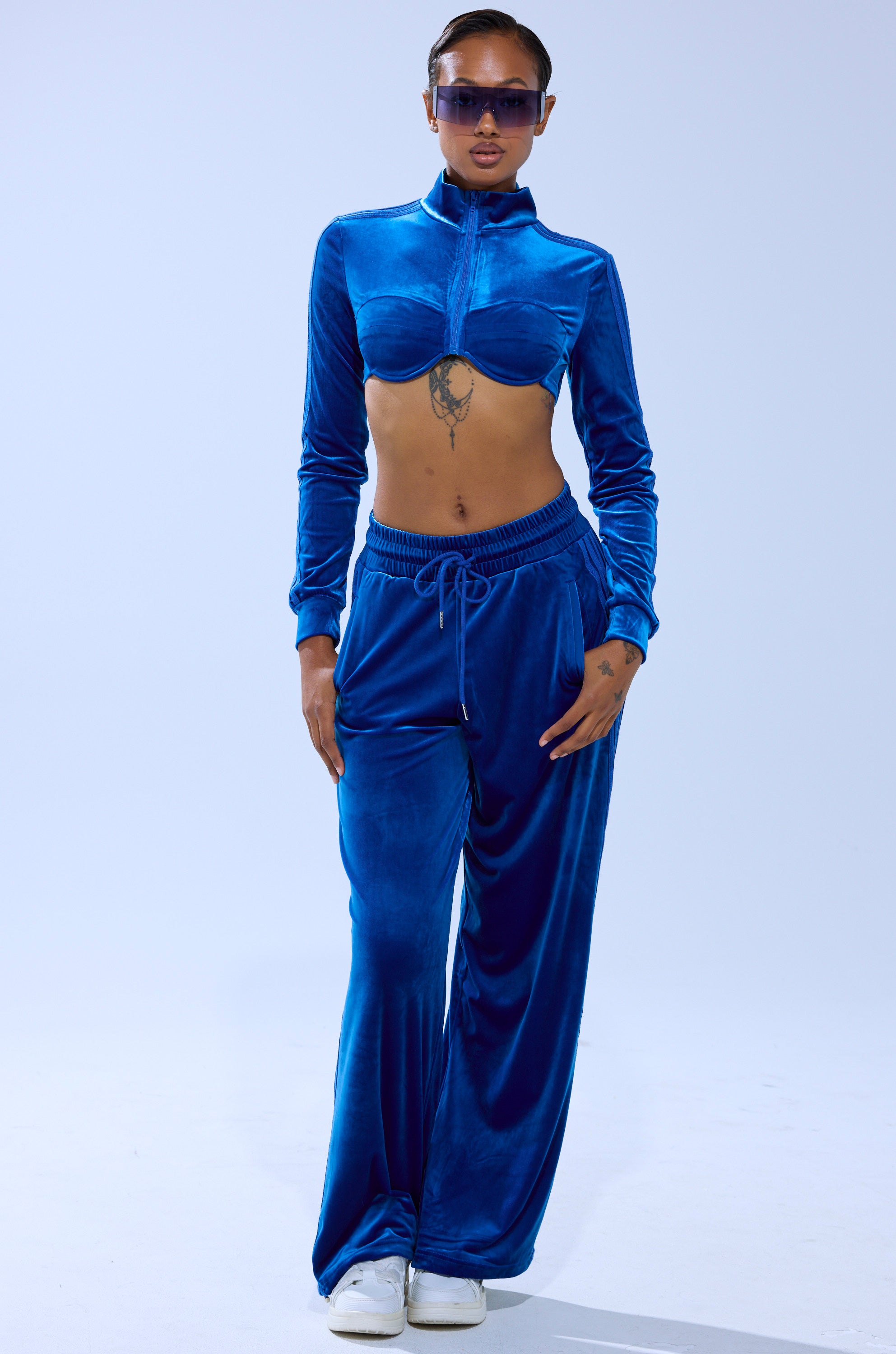 A person poses confidently against a plain light background in the ELECTRIC BLUES CROP ZIP UP SWEATSHIRT and matching wide-leg pants, styled with white sneakers, sunglasses, and hoop earrings.