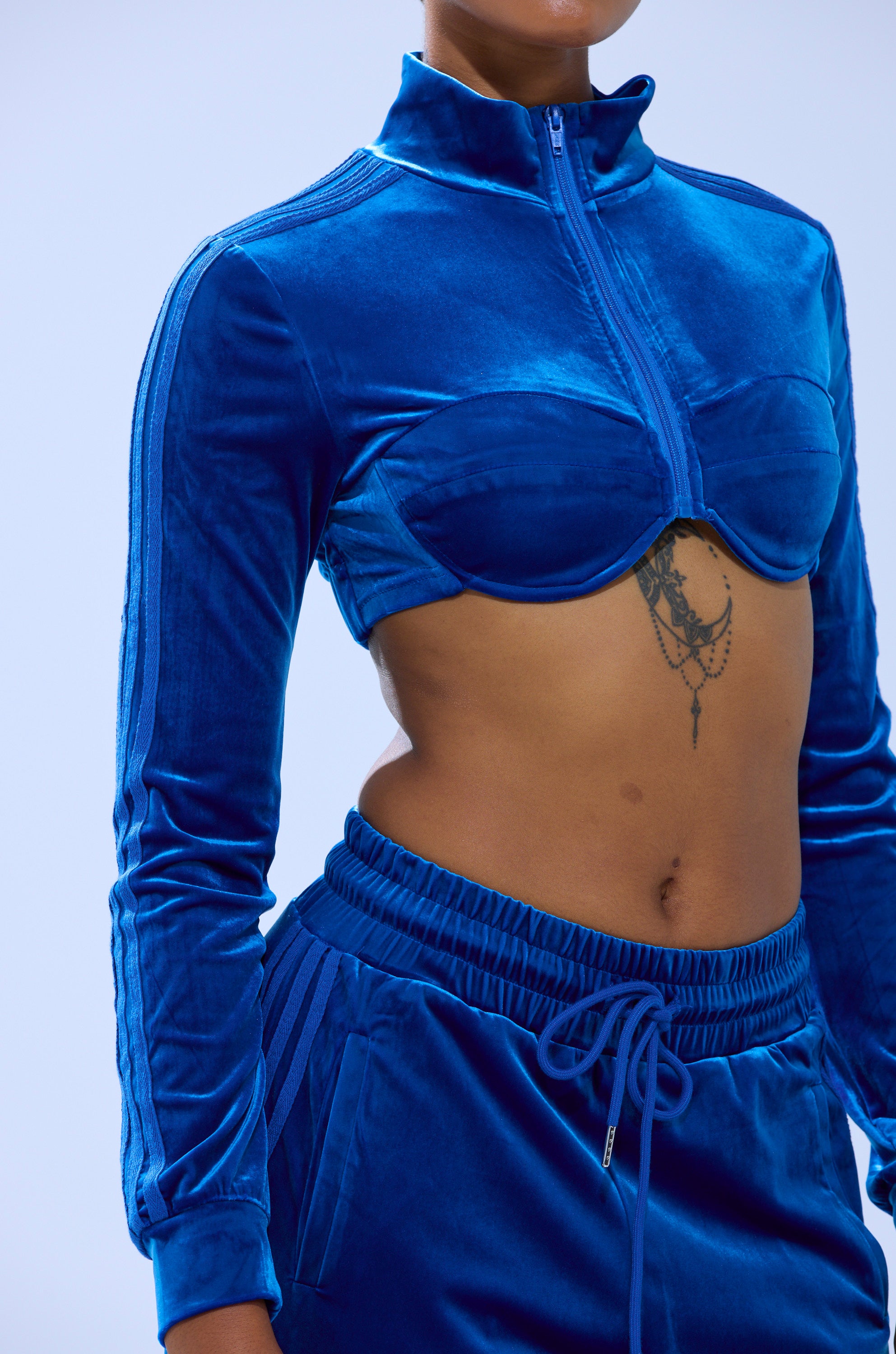 Wearing the ELECTRIC BLUES CROP ZIP UP SWEATSHIRT, a sapphire-blue velour piece with bustier-inspired design, cropped length, and zip front, styled to reveal a tattoo beneath the chest against a light blue background.