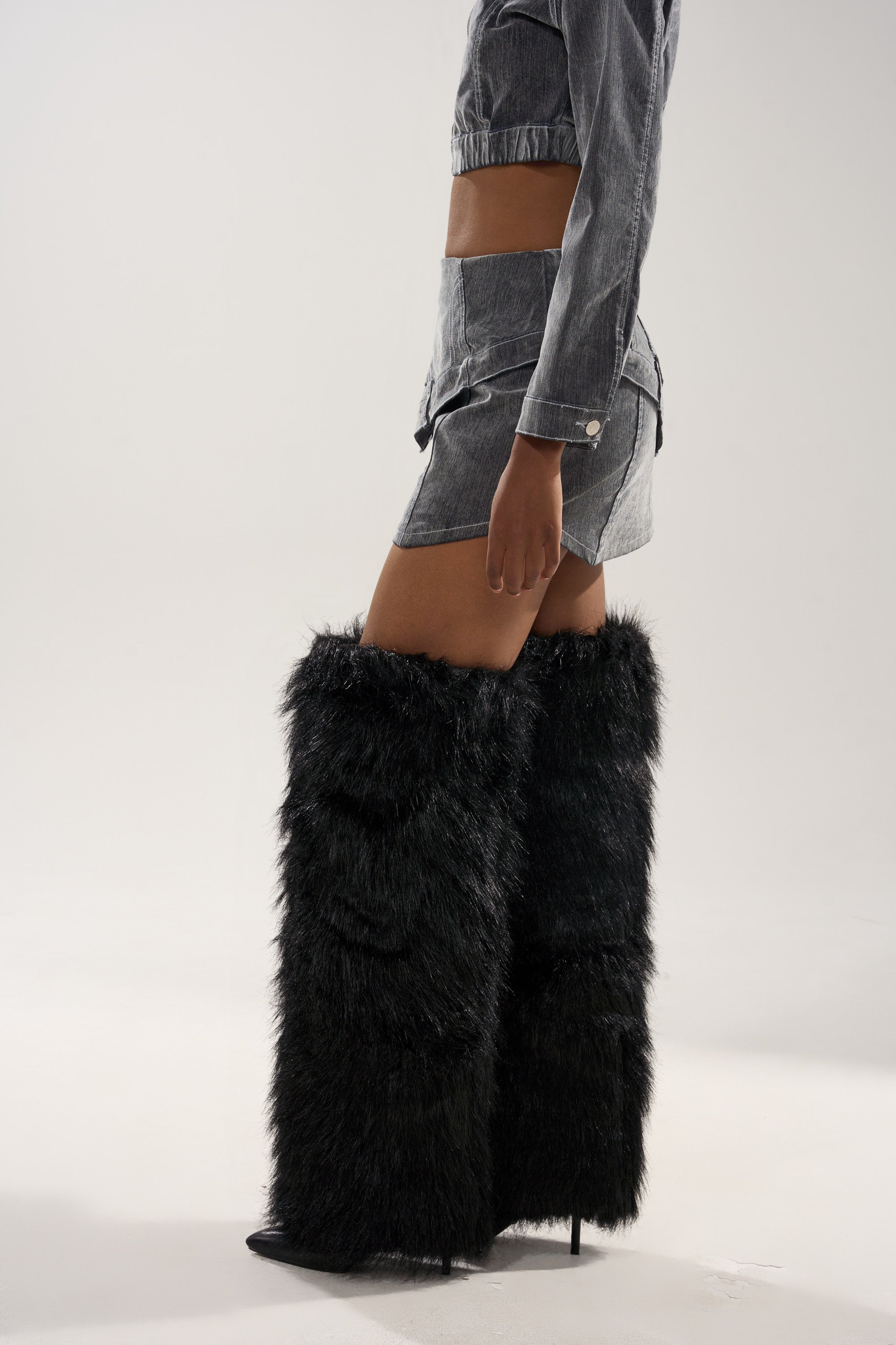 Wearing the ALL FOR LOVE VELVET DENIM MINI SKIRT, a person stands in black furry knee-high boots with pointed toes and high heels, paired with a grey denim jacket against a plain light background.