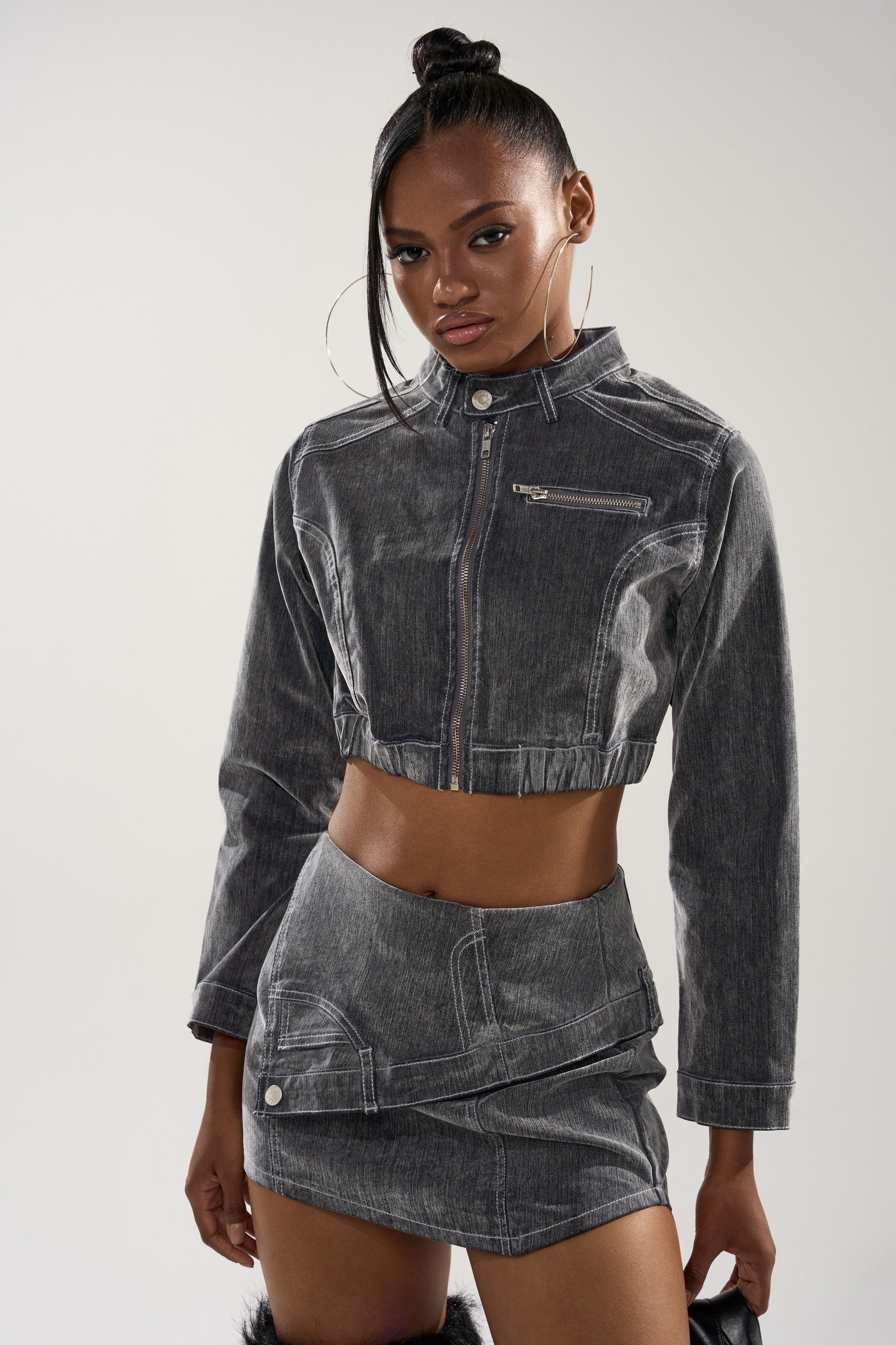 A woman models the ALL FOR LOVE VELVET DENIM MOTO, a vintage grey cropped moto jacket with a chest zipper, paired with a matching gray asymmetrical mini skirt, standing confidently against a plain light background.