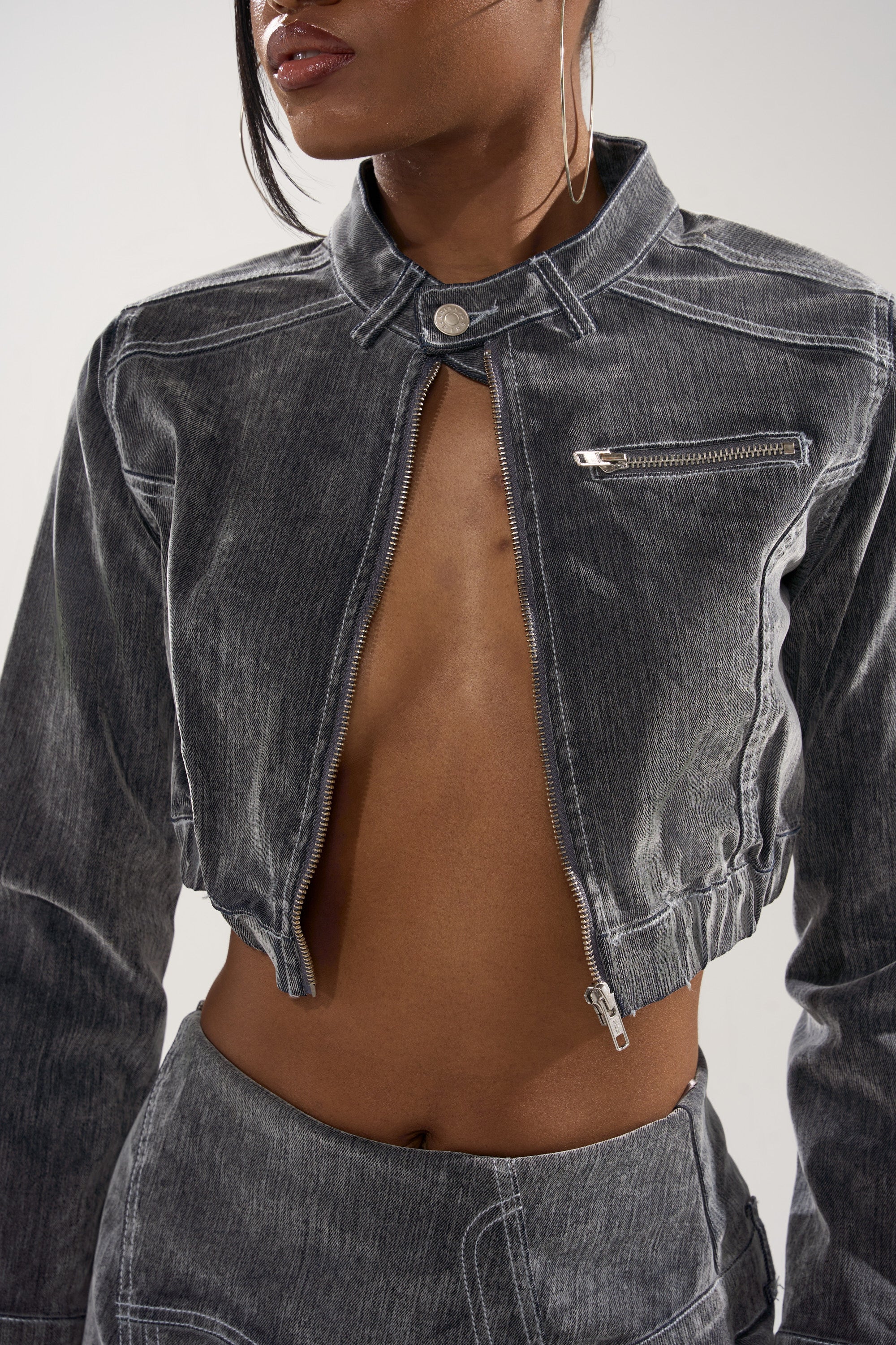 A woman wears the ALL FOR LOVE VELVET DENIM MOTO, a vintage grey wash cropped jacket with a front zipper and chest pocket, paired with matching high-waisted grey pants. Her face is partially out of frame.