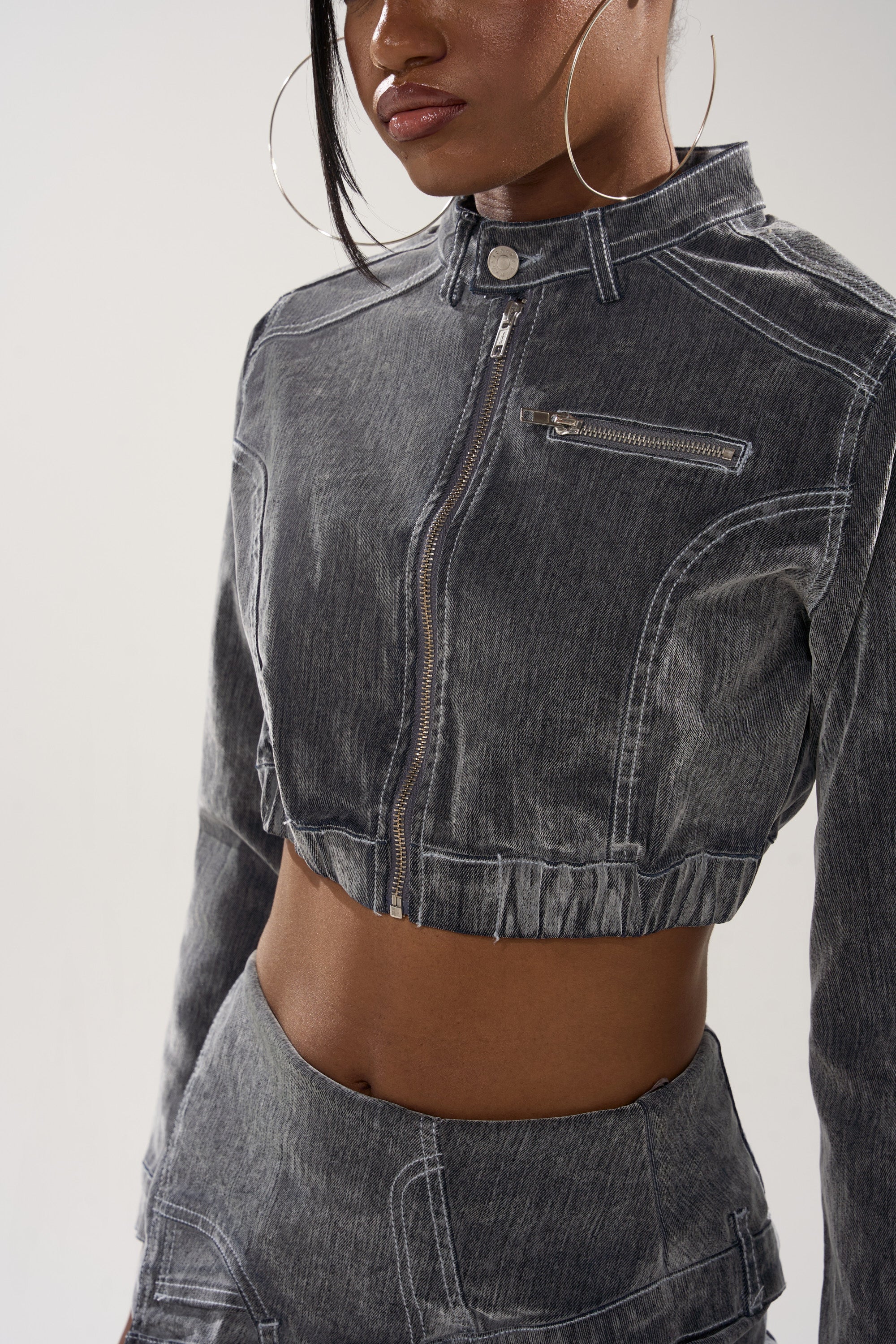 A woman styles the ALL FOR LOVE VELVET DENIM MOTO, a vintage grey wash jacket with white stitching, front zipper, and chest pocket, paired with matching high-waisted pants, large hoop earrings, and straight hair.