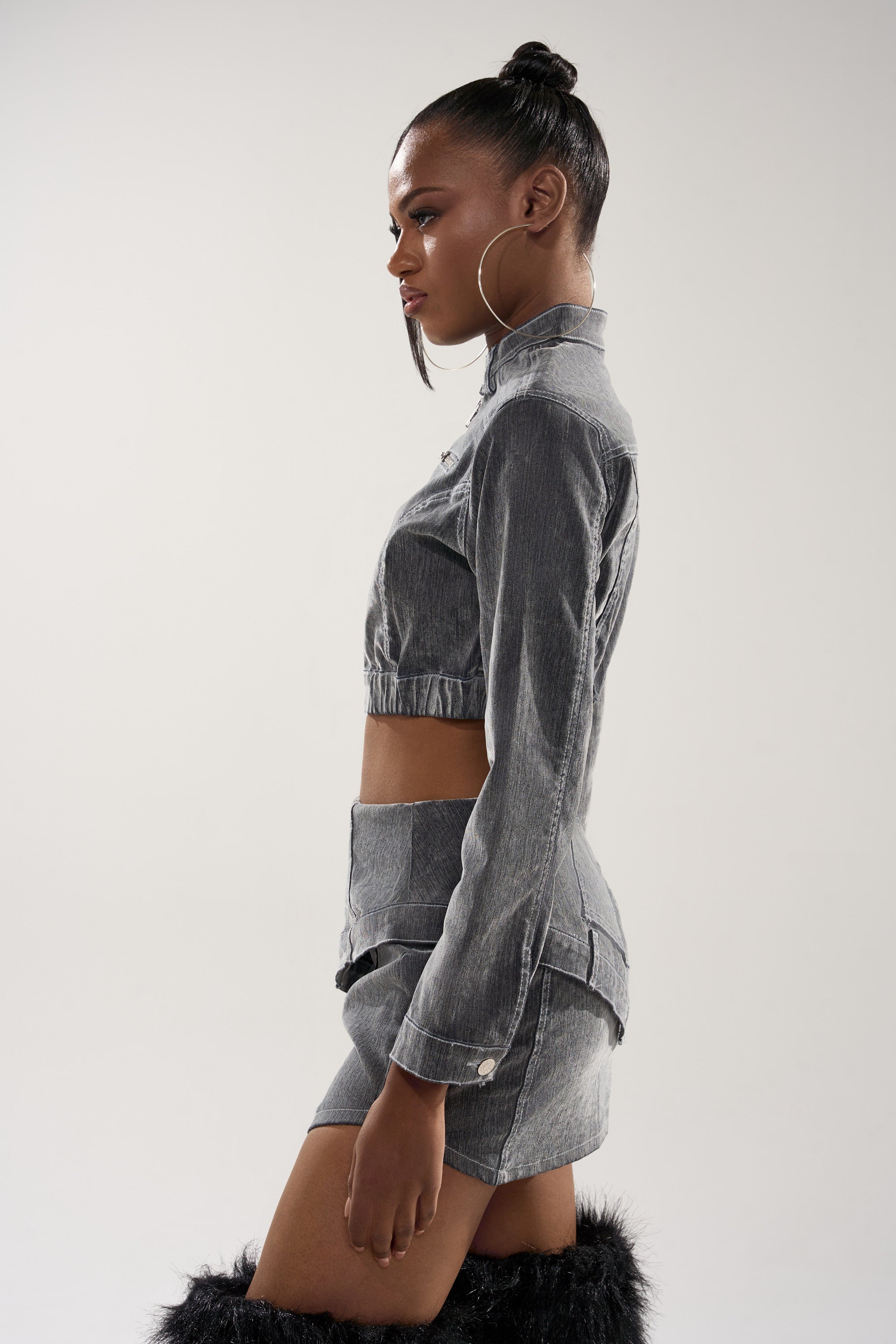 A woman in profile wears the ALL FOR LOVE VELVET DENIM MOTO jacket and matching high-waisted mini skirt, styled with large hoop earrings, a sleek bun, black furry boots, against a plain light background.
