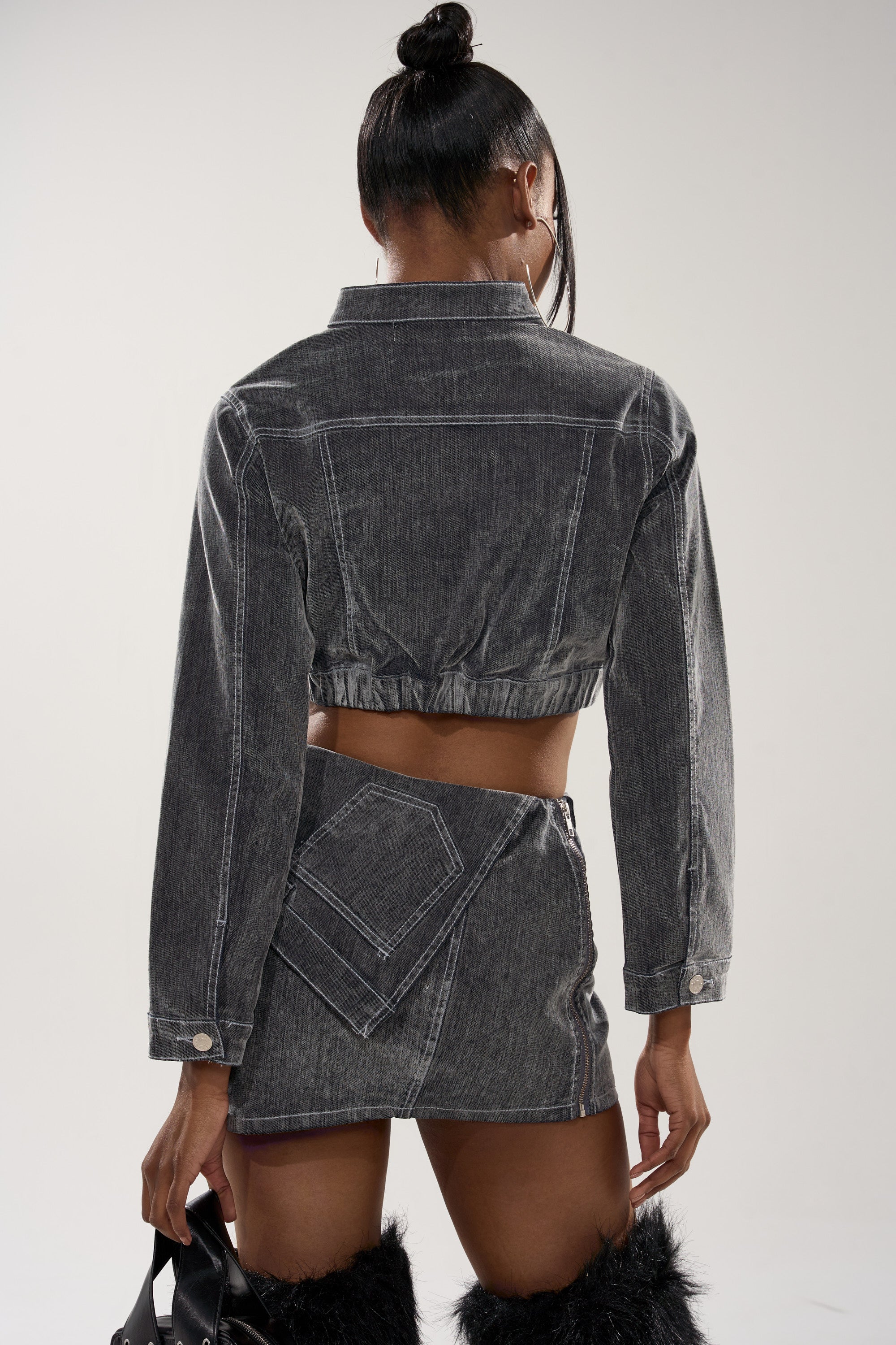 A woman with her hair in a bun faces away, wearing the ALL FOR LOVE VELVET DENIM MOTO jacket and matching high-waisted mini skirt with large back pockets, furry black boots, and holding a black handbag.
