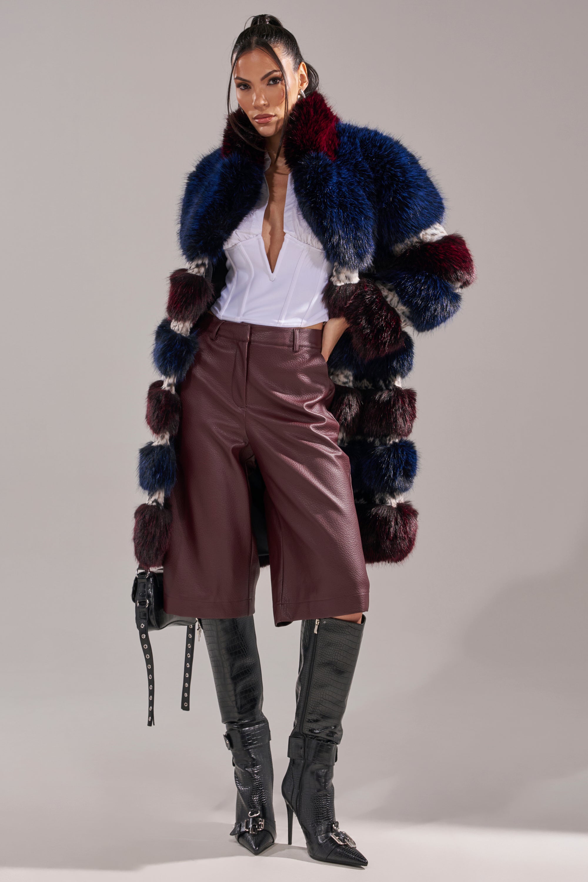 A woman stands confidently against a plain background in a colorful AKIRA Label faux fur coat over a white top, burgundy BEST ONE YET BERMUDA SHORT high-rise faux leather shorts, black knee-high boots, and holding a small black bag. 9