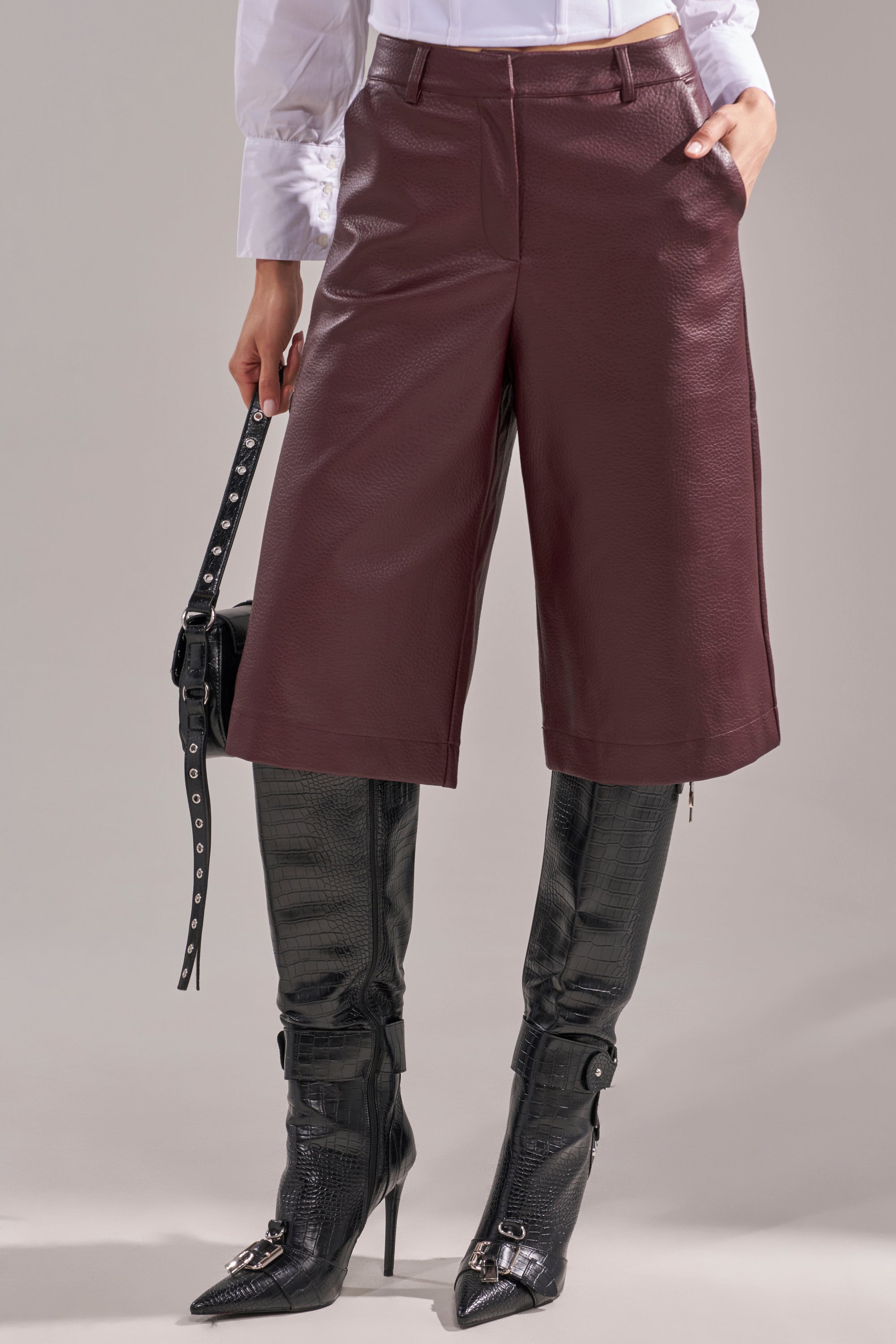 A person wears a white blouse, BEST ONE YET BERMUDA SHORT in burgundy faux leather, black pointed-toe high-heeled boots, and holds a black bag with silver buckles. The background is plain gray. 1