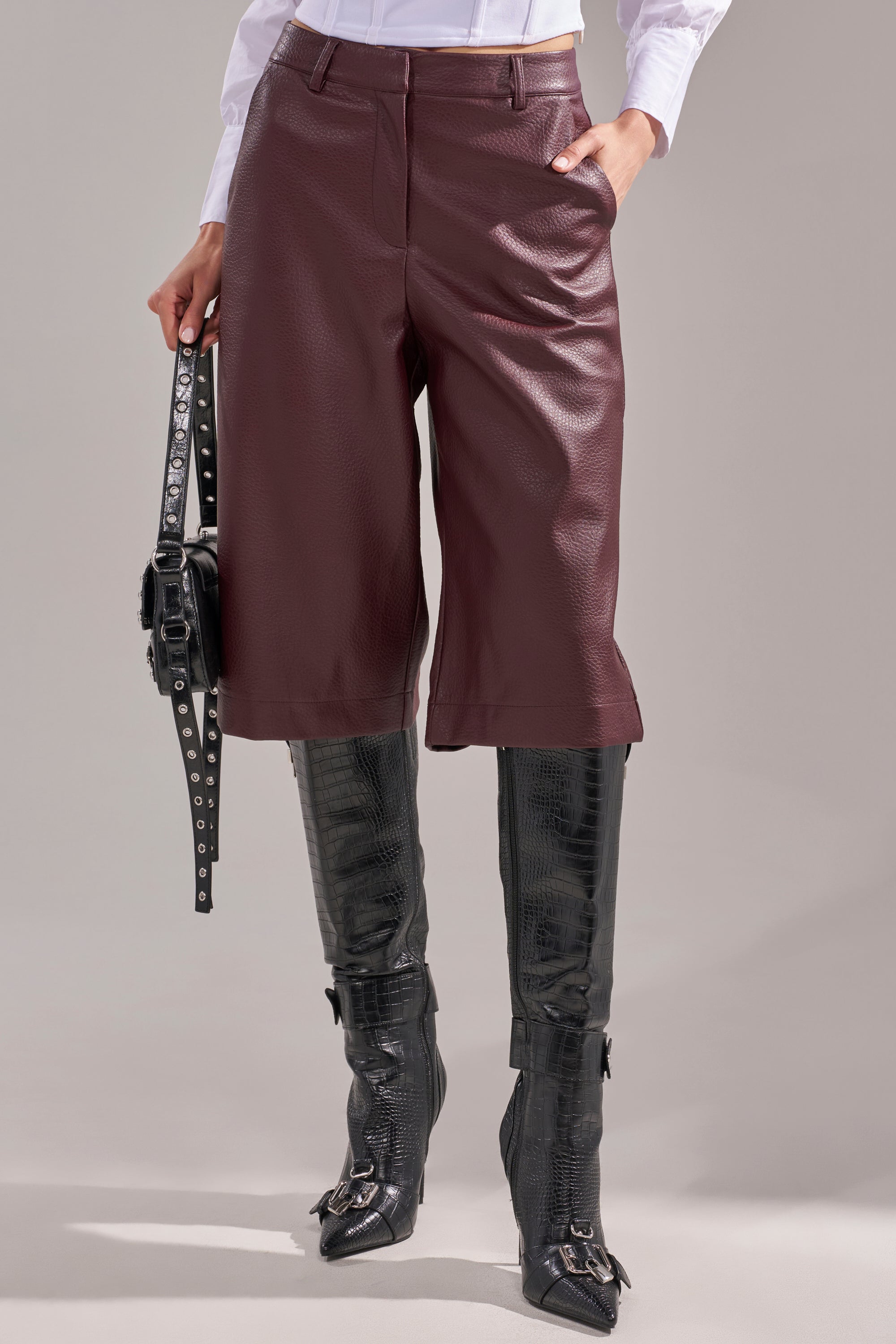 A person wearing BEST ONE YET BERMUDA SHORT in burgundy faux leather, a white shirt, tall black textured boots, and holding a black studded handbag by their side. 2