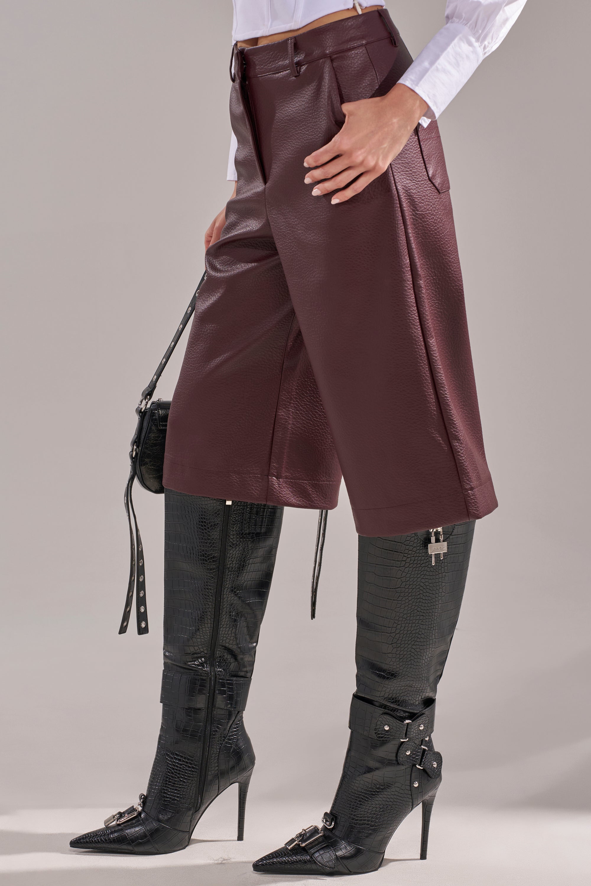 A person wearing BEST ONE YET BERMUDA SHORT faux leather shorts in burgundy, a white long-sleeve shirt, black knee-high heeled boots with buckles, and holding a black handbag at their side. 3