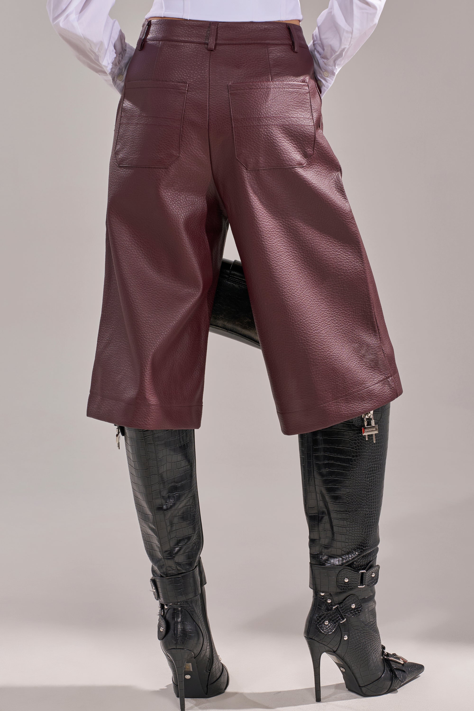 Wearing the BEST ONE YET BERMUDA SHORT in burgundy faux leather and black knee-high heeled boots with straps and buckles, a person stands hands on hips, photographed from behind. 4