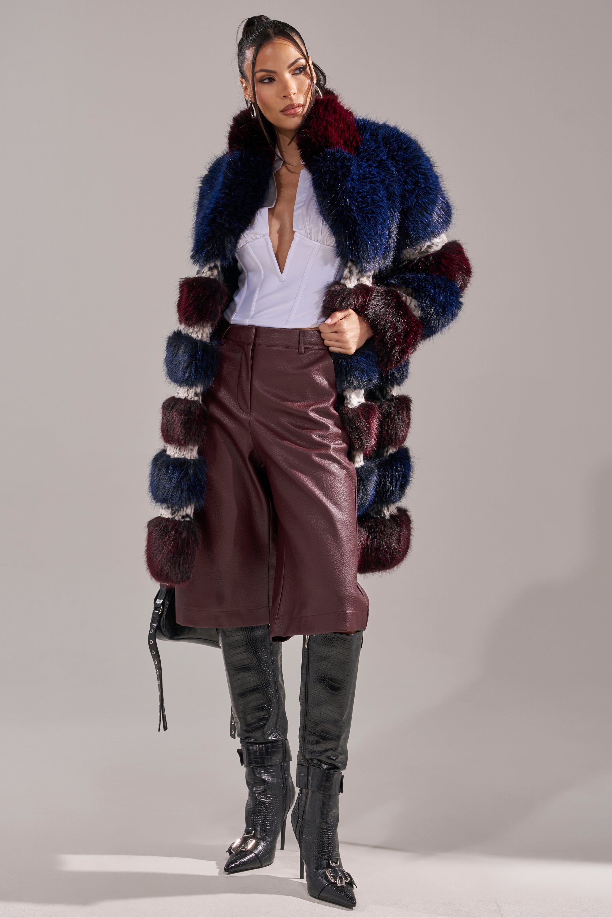 A woman wearing an AKIRA Label white blouse, BEST ONE YET BERMUDA SHORT, tall black boots, and a fluffy striped fur coat in burgundy and navy poses confidently against a plain background. 8