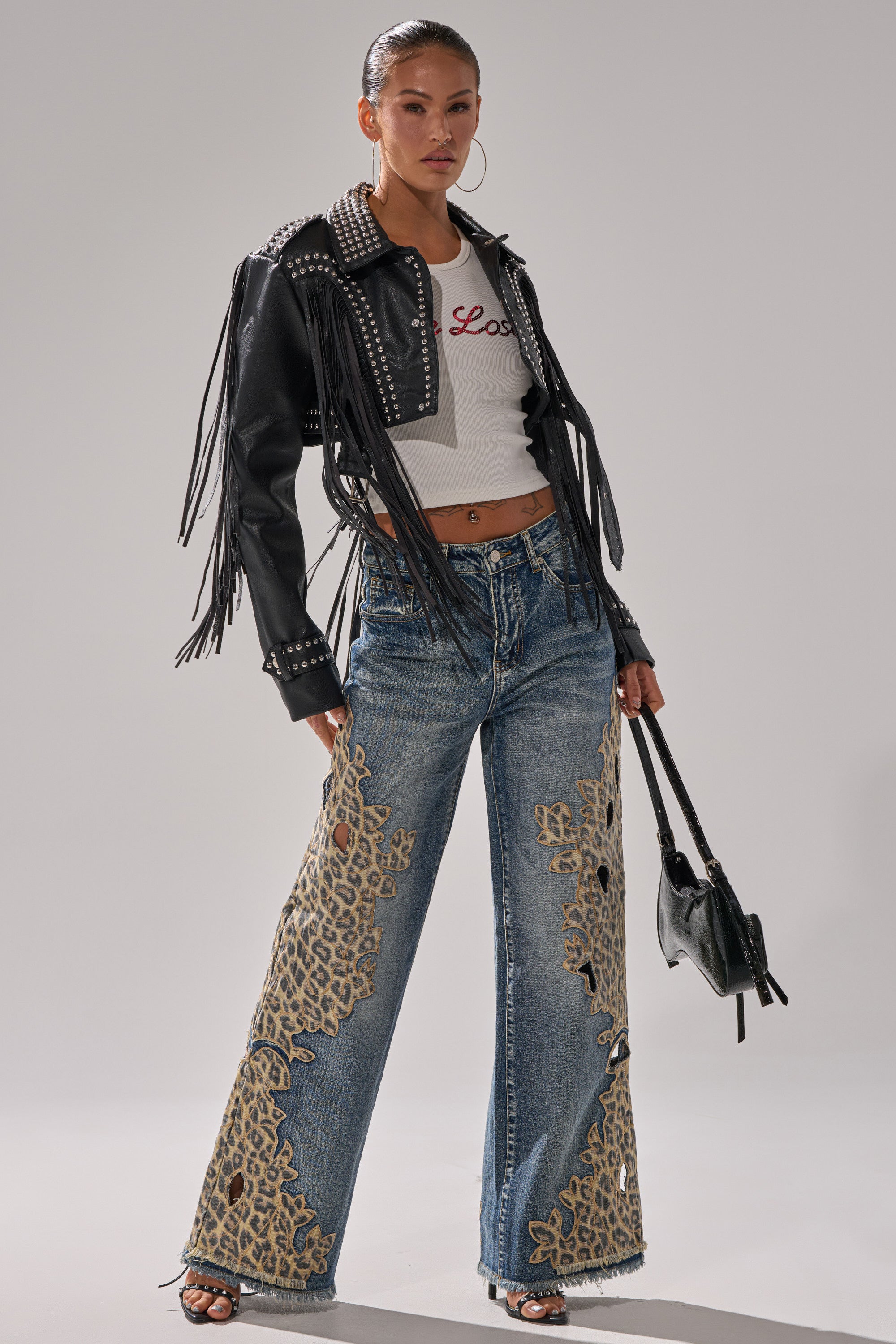 A woman poses in cheetah print wide-leg pants, specifically the AMIGAS CHEETAH PRINT WIDE LEG PANT, styled with a studded black leather jacket, white crop top, black handbag, hoop earrings, and strappy black sandals.
