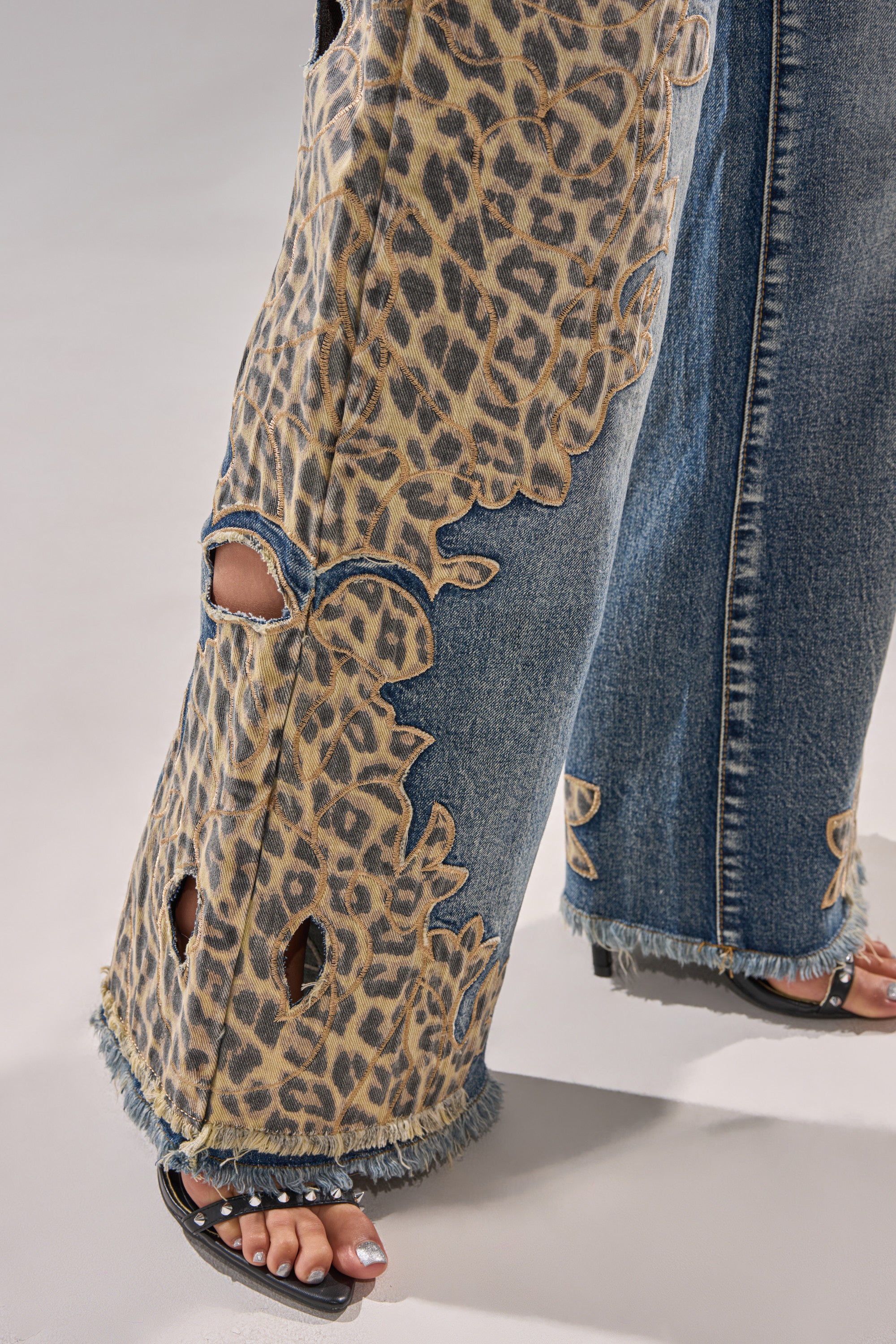 Close-up of AMIGAS CHEETAH PRINT WIDE LEG PANT featuring frayed hems, cheetah print patches, and cut-out details, paired with black sandals on a light background.