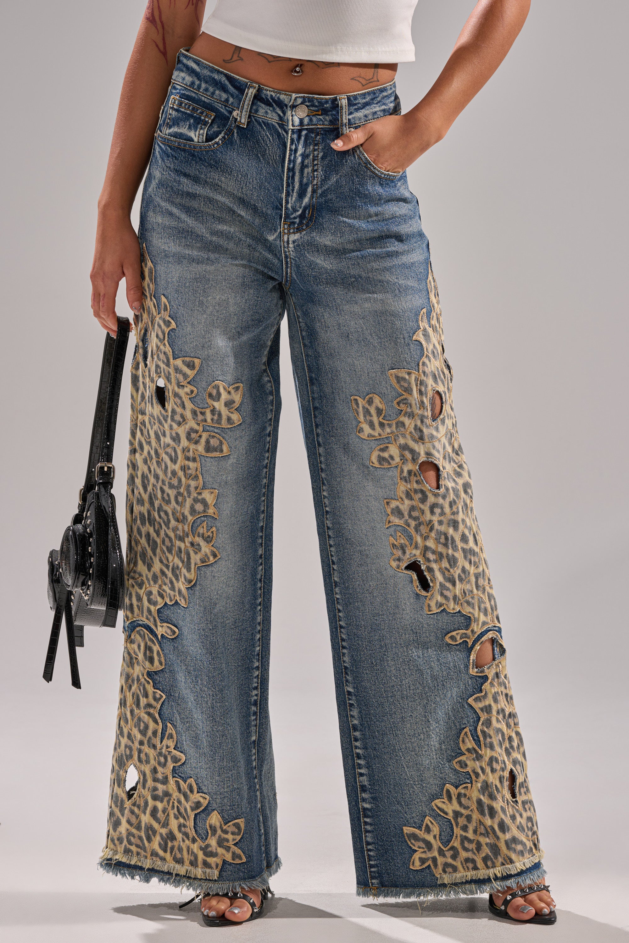 A person wears AMIGAS CHEETAH PRINT WIDE LEG PANT featuring cheetah print, wide legs, and frayed cut-outs, paired with a white crop top and a small black handbag. The image is cropped from the shoulders down.