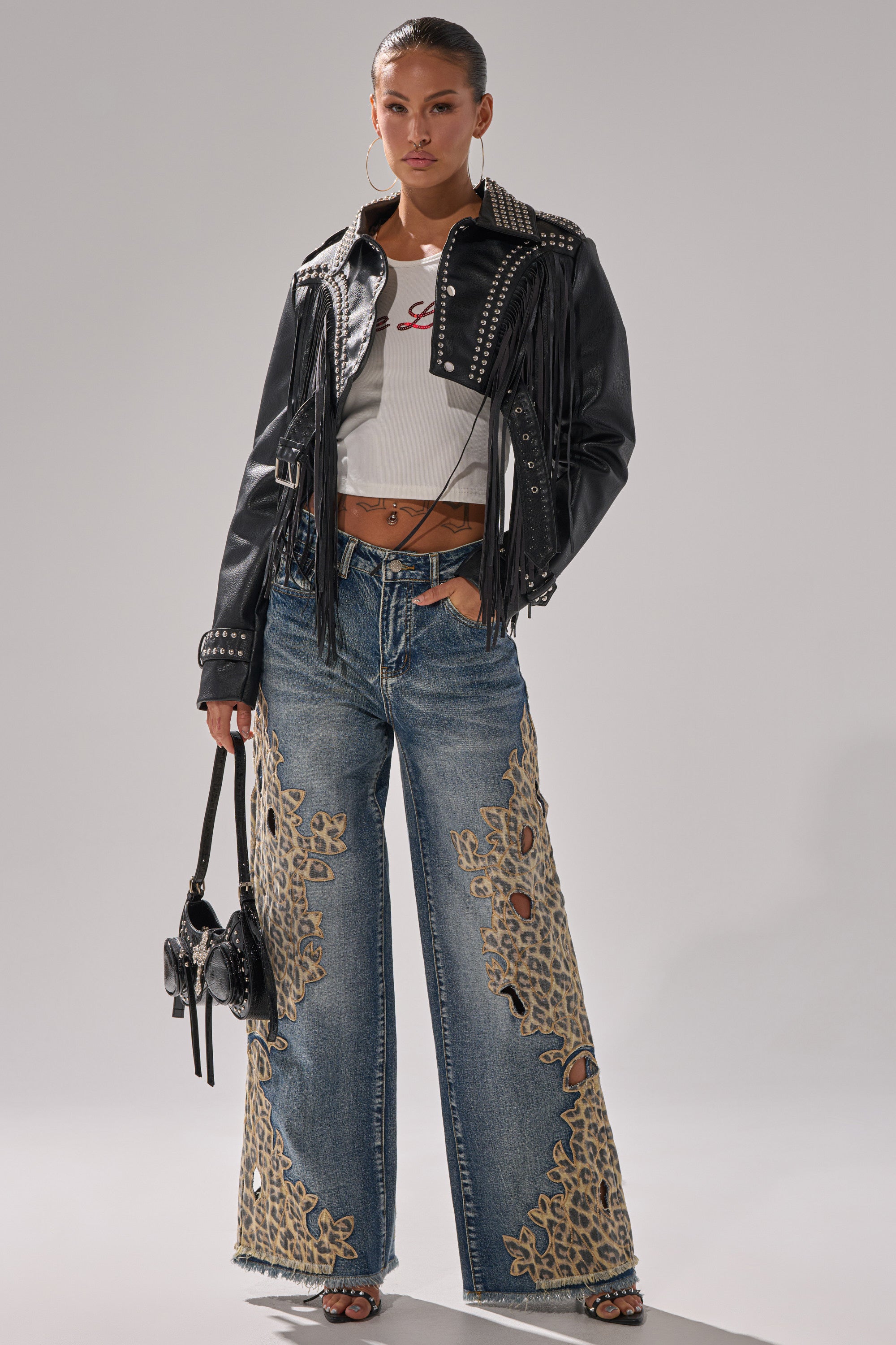 A woman poses in cheetah print wide leg pants, a white crop top, studded black leather jacket with fringe, heeled sandals, and a small black handbag against a plain background.