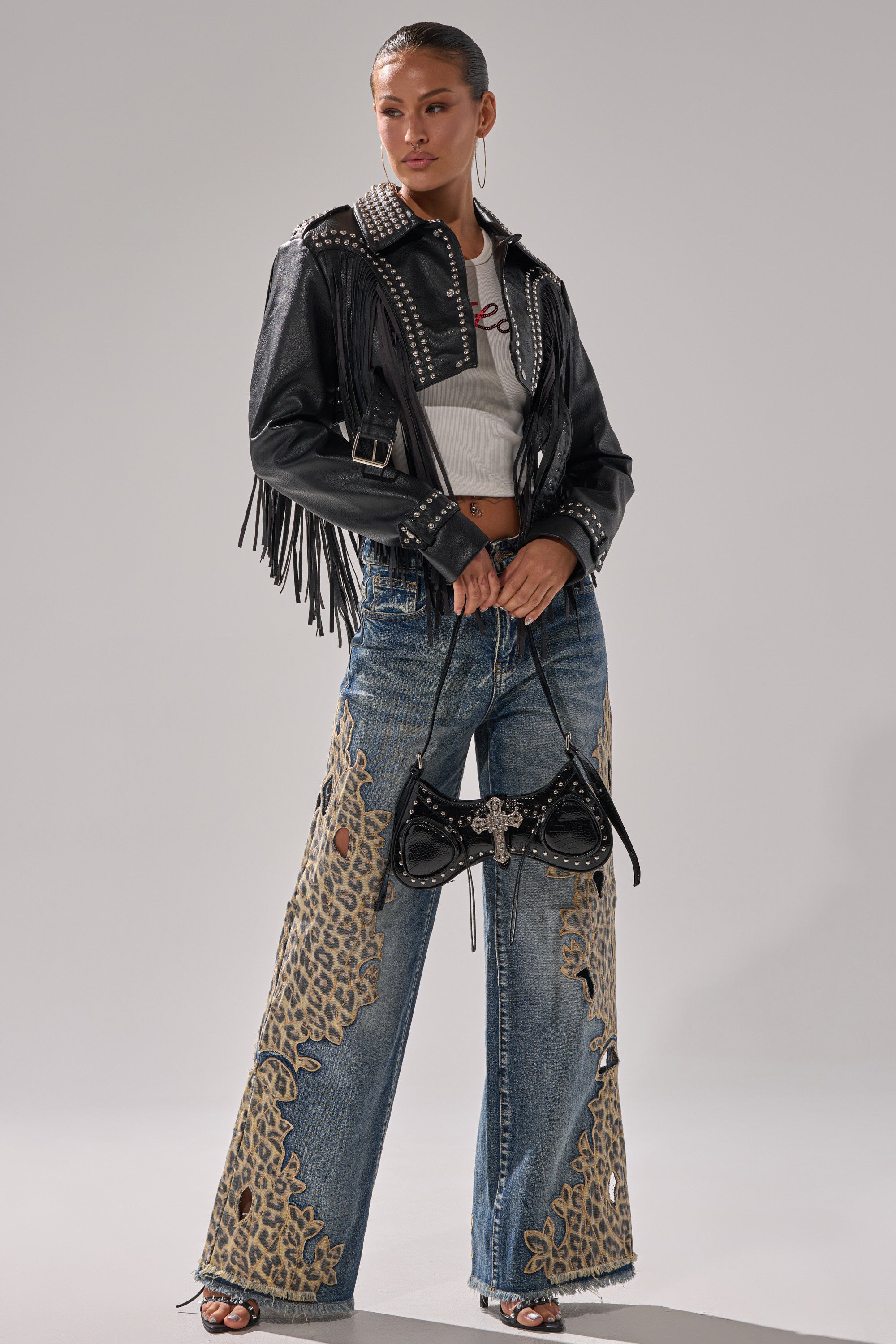 A woman stands confidently in a black leather fringe jacket, white crop top, AMIGAS CHEETAH PRINT WIDE LEG PANT, black sandals, and holds a black studded handbag.