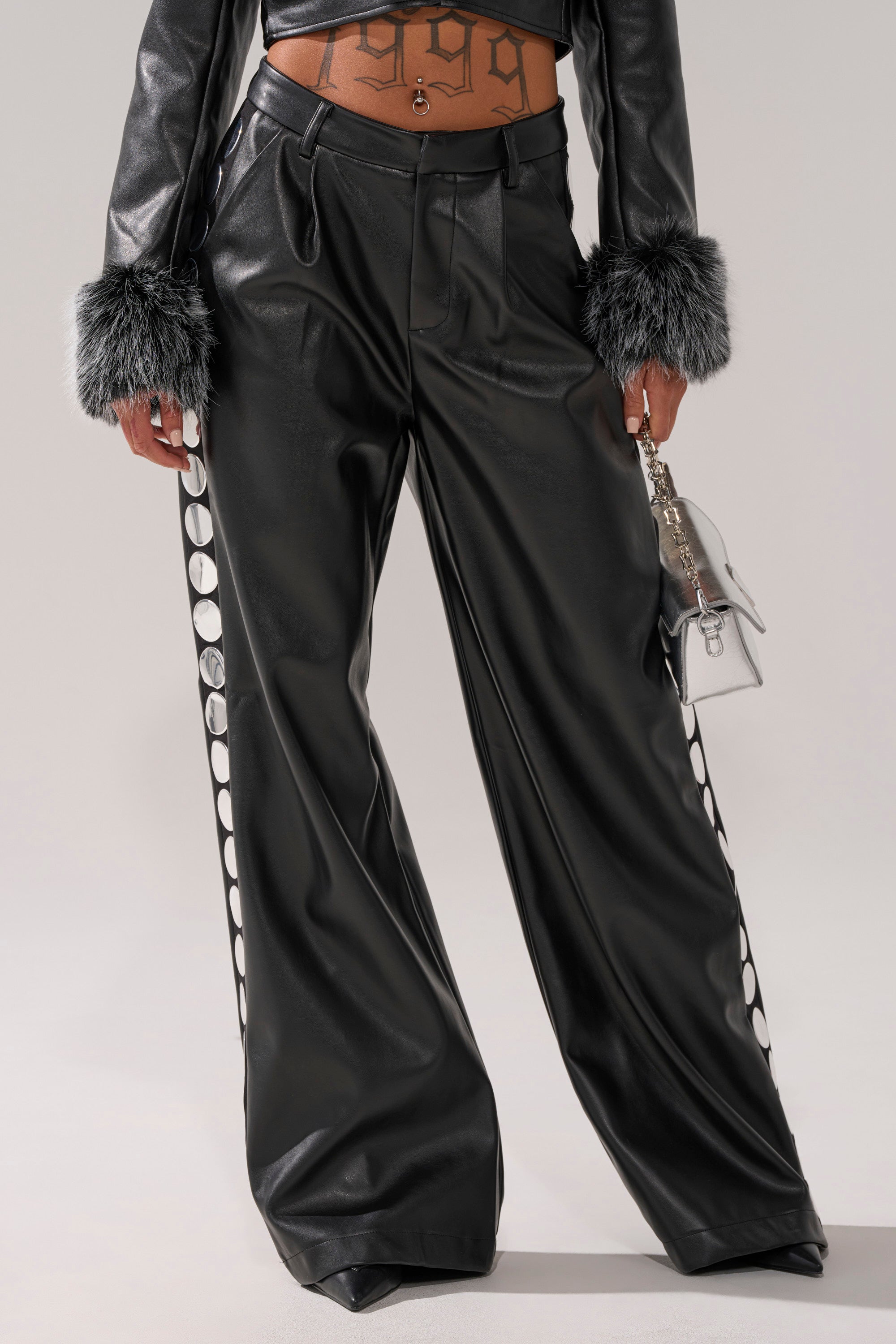 Wearing the TIME AFTER TIME FAUX LEATHER TROUSER—black high-rise, wide-leg pants with silver grommet accents—paired with a cropped leather jacket with faux fur cuffs and holding a small silver handbag. Only lower torso and legs are visible.