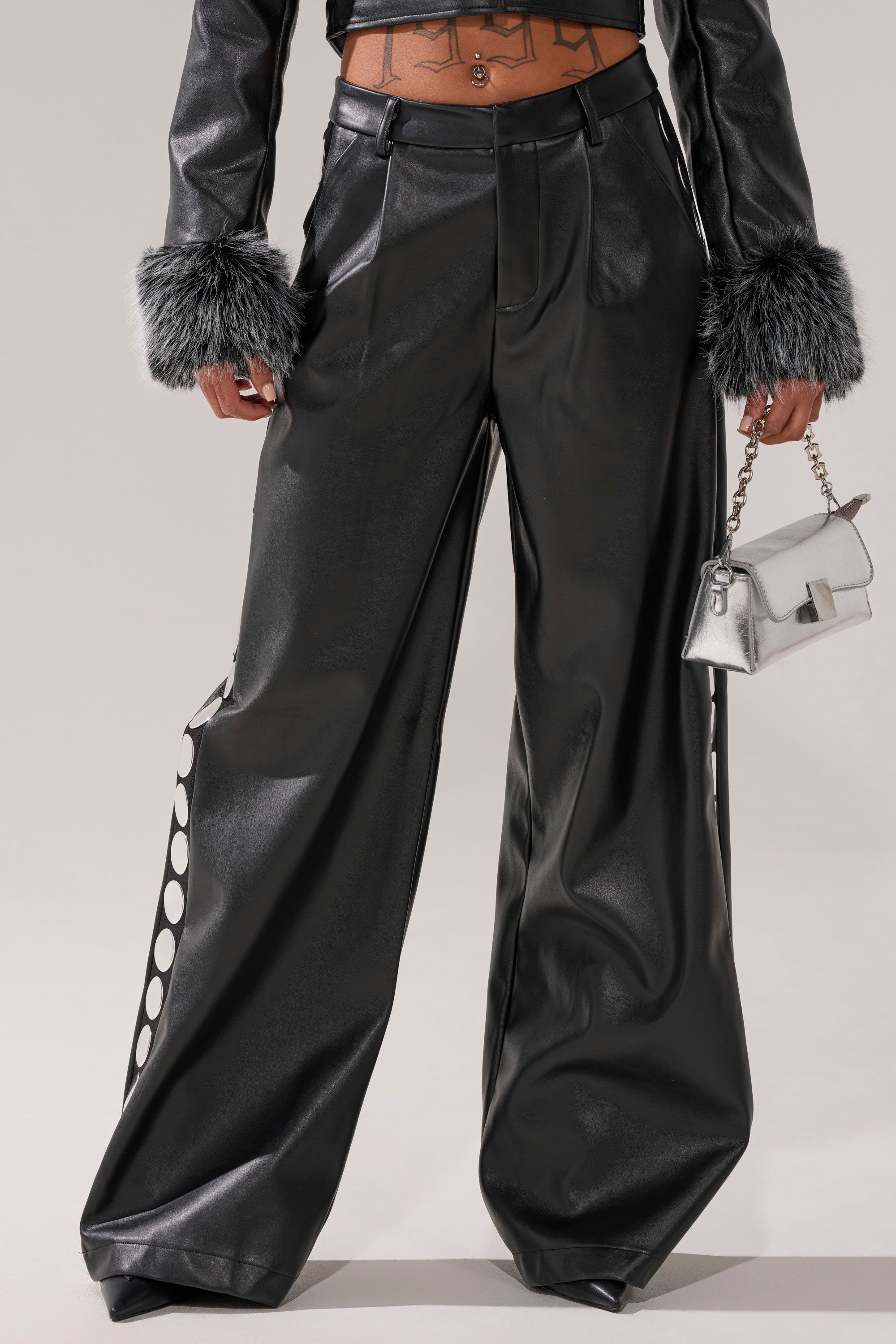 Wearing the TIME AFTER TIME FAUX LEATHER TROUSER—high-rise wide-leg vegan leather pants with white polka dot side stripes—a matching black jacket with gray faux fur cuffs, and a small metallic silver handbag.