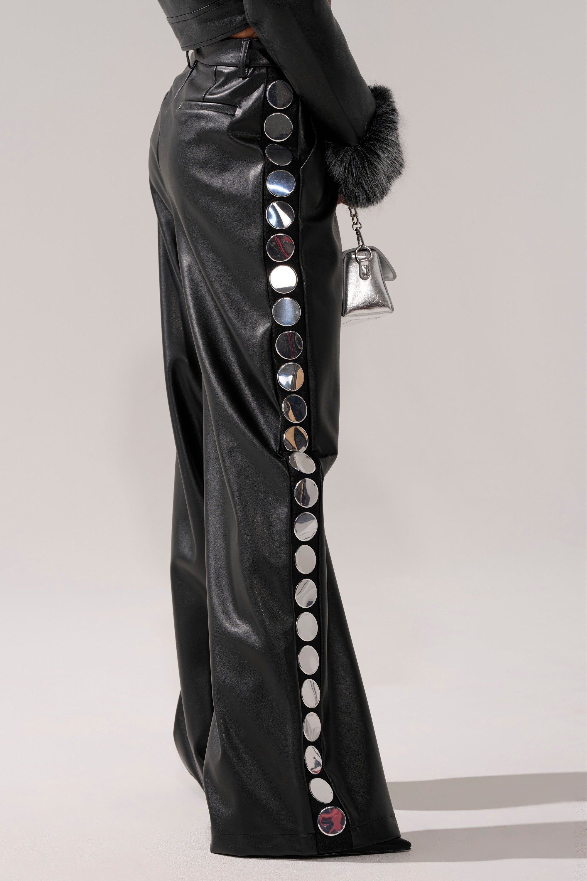 A person wears the TIME AFTER TIME FAUX LEATHER TROUSER—black vegan leather pants with large silver circular embellishments down the side—while holding a small metallic handbag with a black faux fur cuff, lower torso and legs visible.