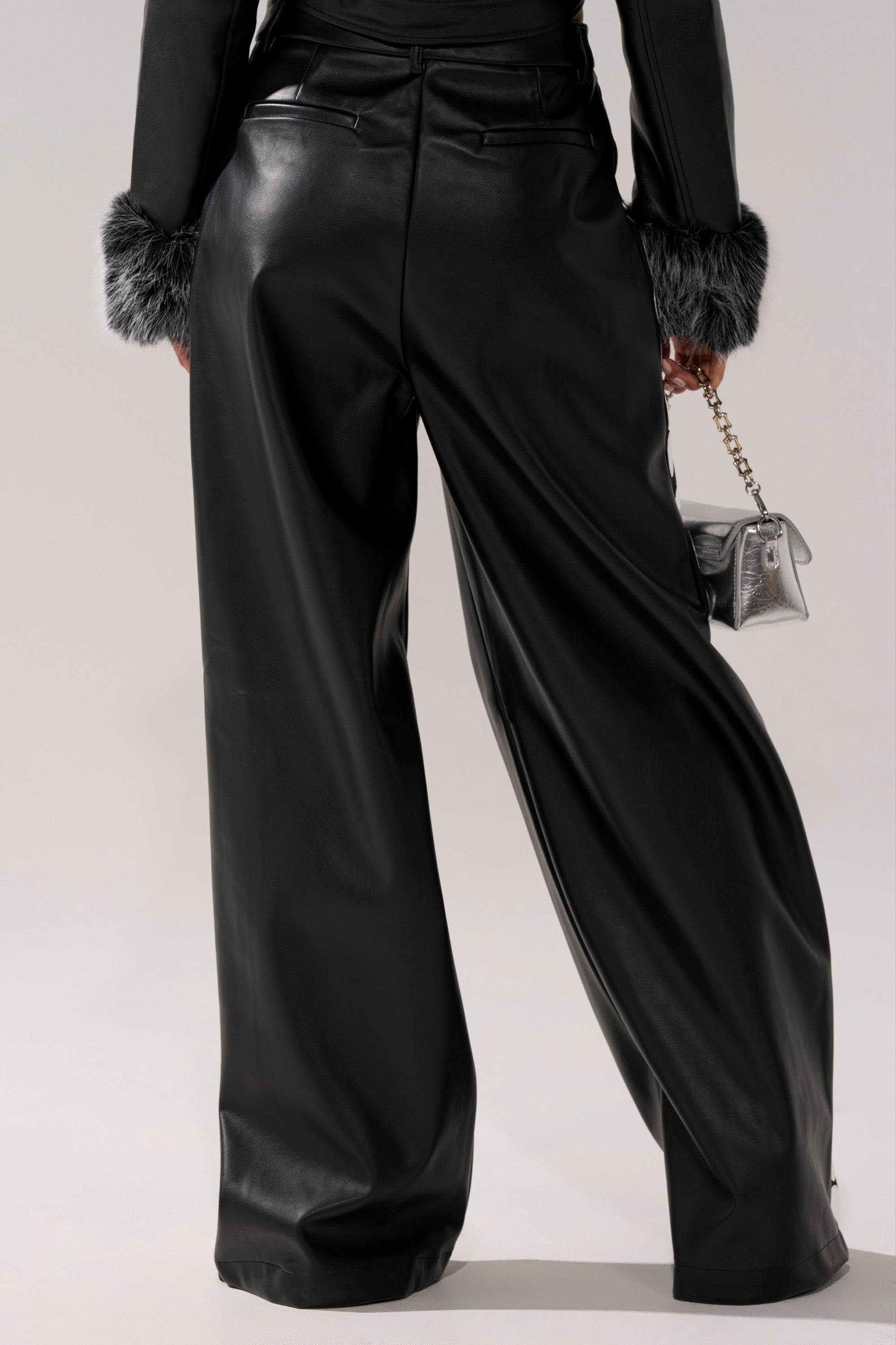 A person wearing the TIME AFTER TIME FAUX LEATHER TROUSER and a coordinating faux fur-cuffed jacket holds a small silver handbag, pictured from the back.