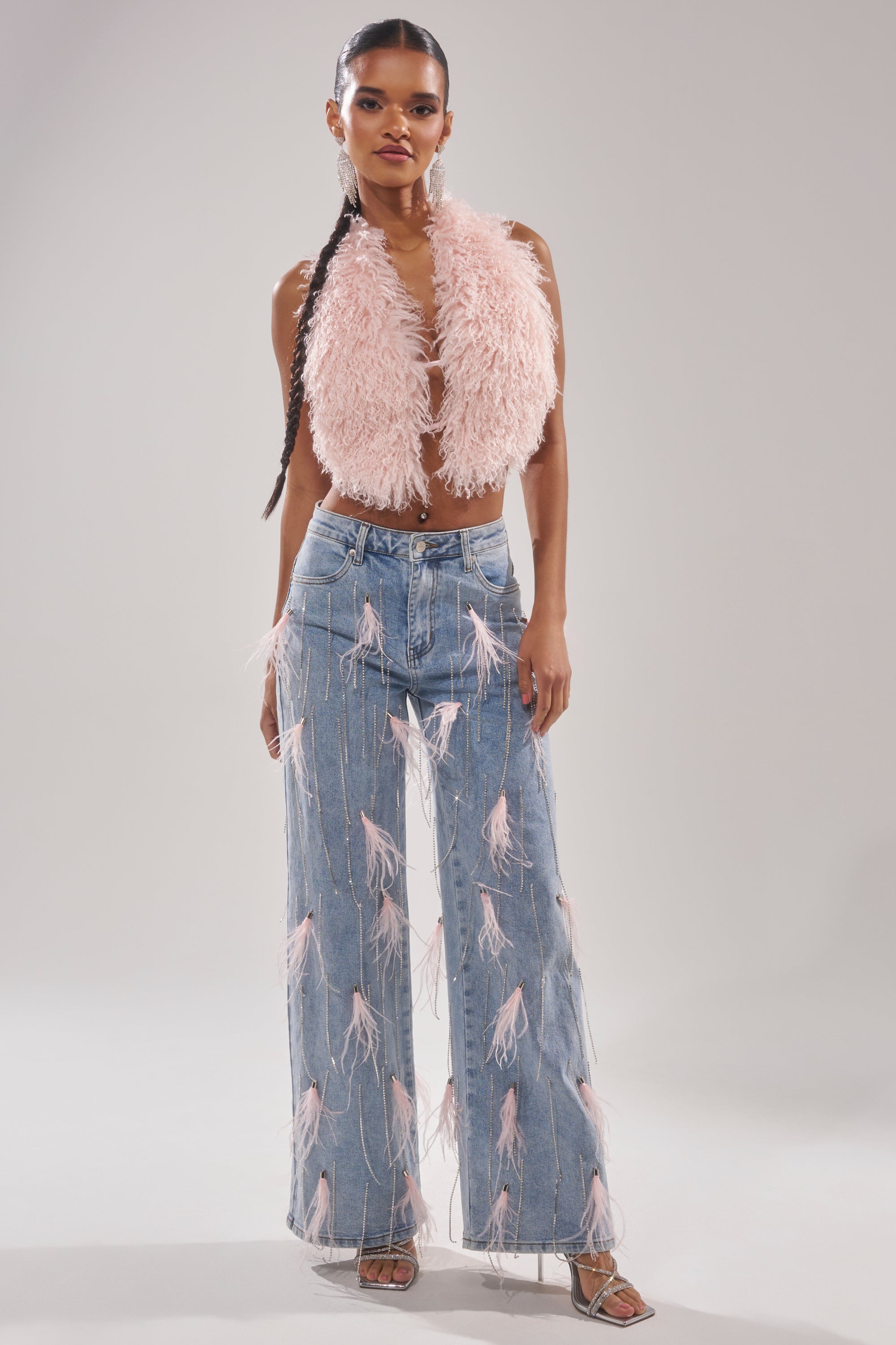 A woman wears the SHAKE IT FOR ME GIRL Pink Feather Embellished Denim high-waisted jeans with pink fringe, paired with a pink fuzzy sleeveless crop top and strappy heels. The backdrop is plain light gray. 1