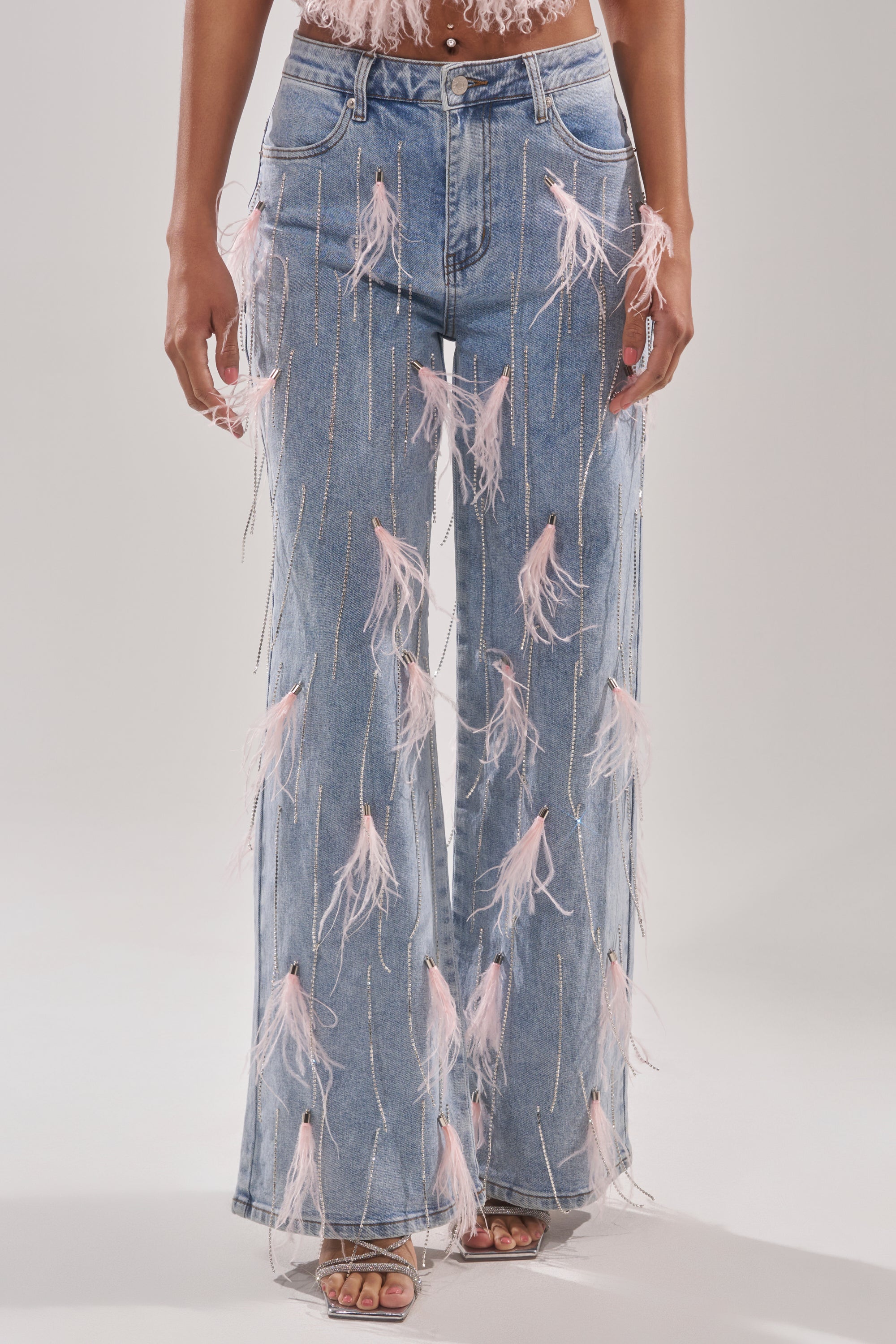 A person stands against a plain light background wearing SHAKE IT FOR ME GIRL PINK FEATHER EMBELLISHED DENIM—light blue wide-leg jeans with pink feathers and silver rhinestone chains—paired with open-toe heels. 0