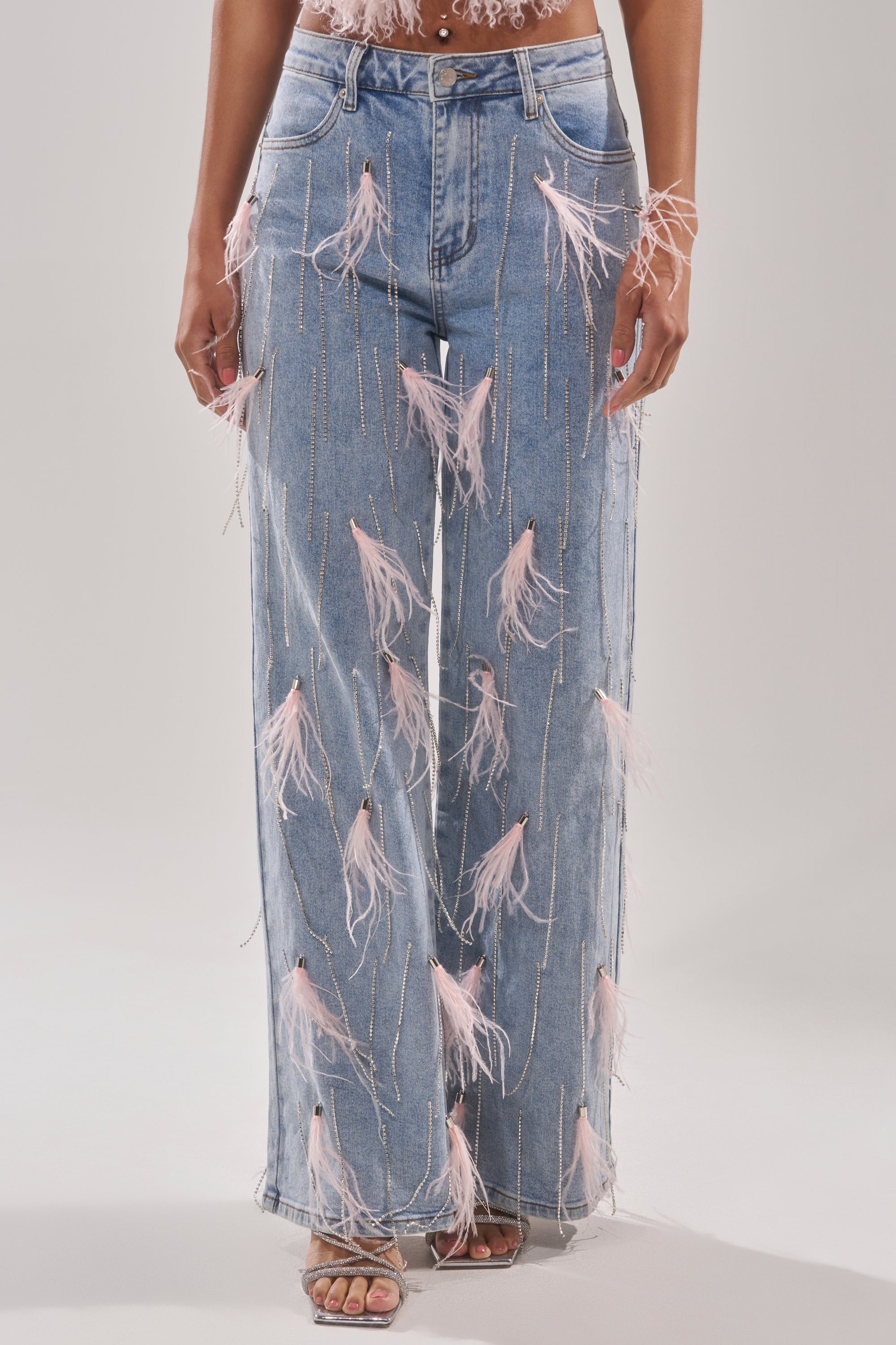 Wearing the SHAKE IT FOR ME GIRL pink feather embellished denim—high-waisted, wide-leg jeans with feather accents—paired with open-toe heeled sandals and a top revealing the midriff. 2