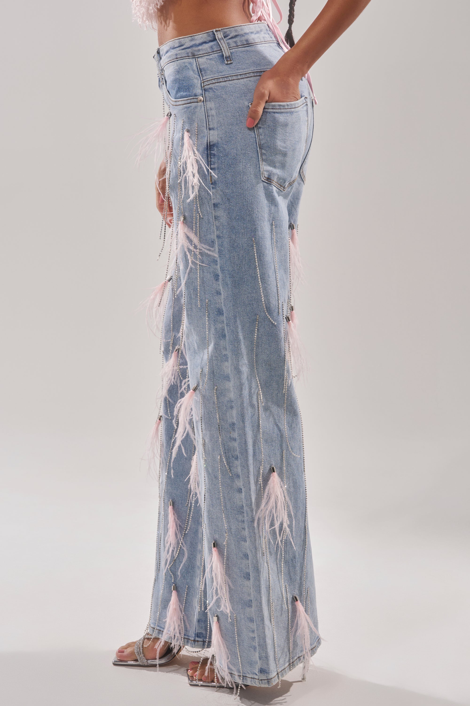 Wearing the SHAKE IT FOR ME GIRL PINK FEATHER EMBELLISHED DENIM with chains and rhinestones, paired with open-toed heels, a person stands against a plain light background. 3