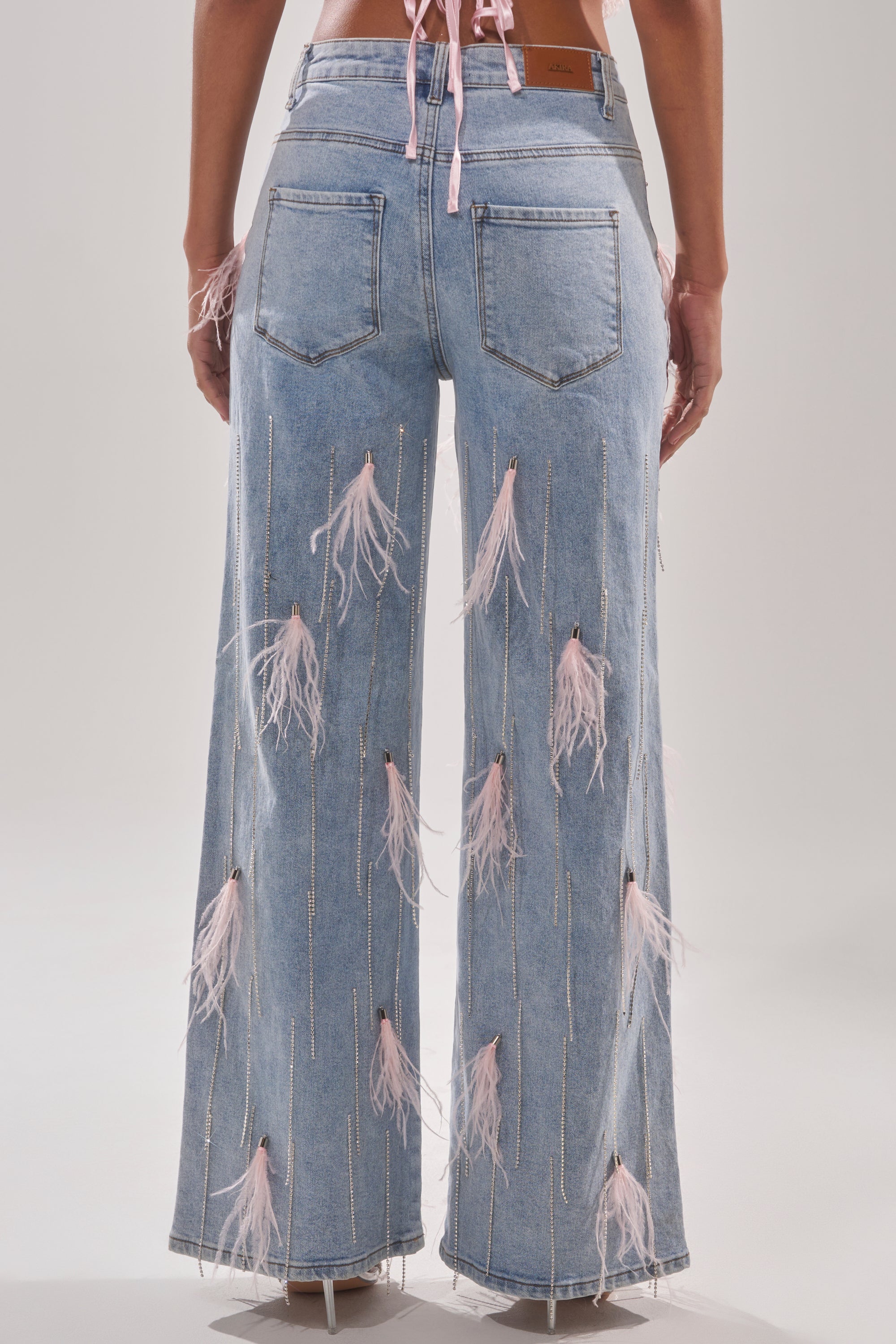 Back view of a person wearing the SHAKE IT FOR ME GIRL PINK FEATHER EMBELLISHED DENIM, featuring light blue denim adorned with scattered pink feathers and vertical glittering accents, paired with transparent high-heeled shoes. 4