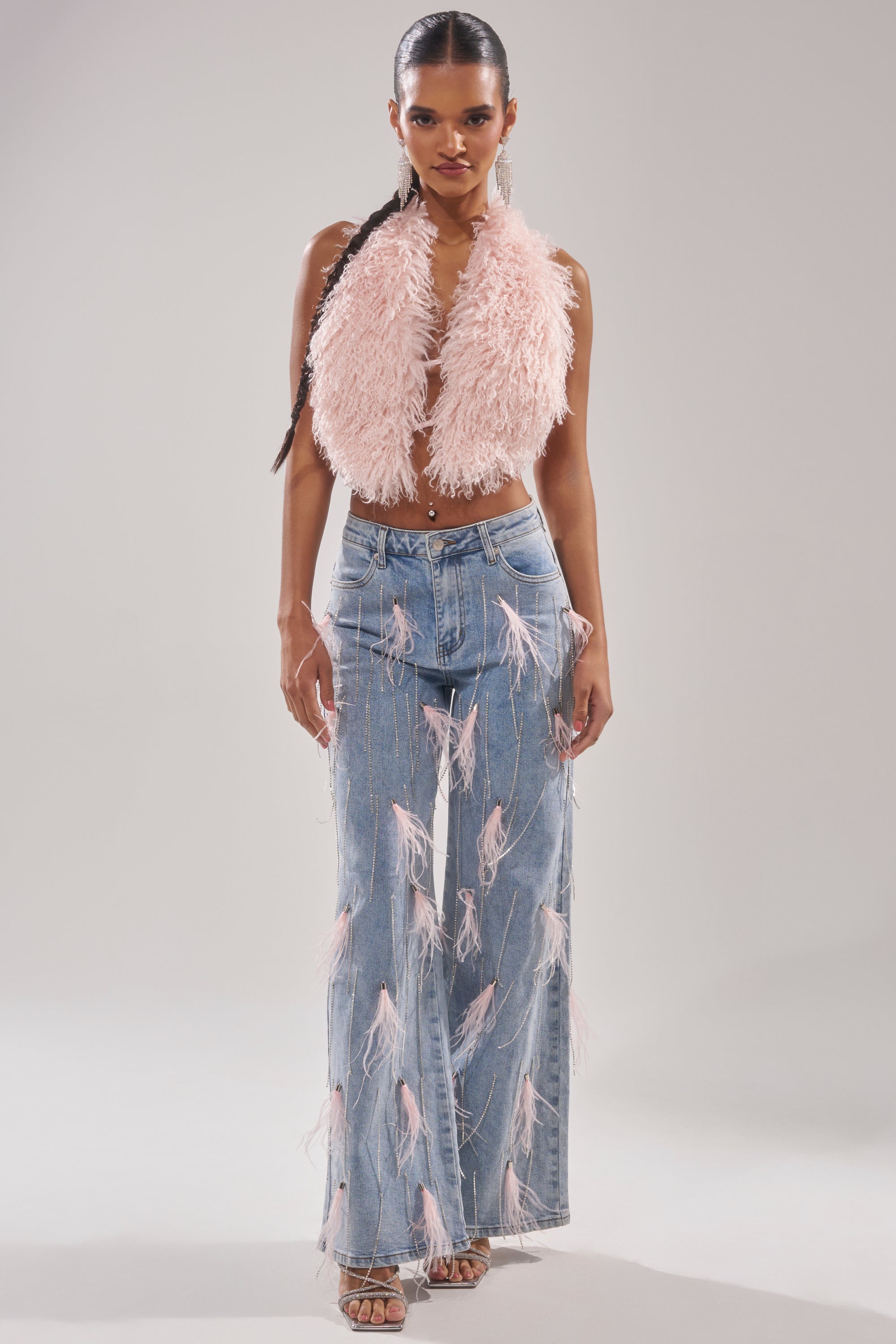 A woman in a sleeveless, pale pink fuzzy crop top and SHAKE IT FOR ME GIRL PINK FEATHER EMBELLISHED DENIM poses forward, her long braid and open-toe heels completing the look against a plain background. 5