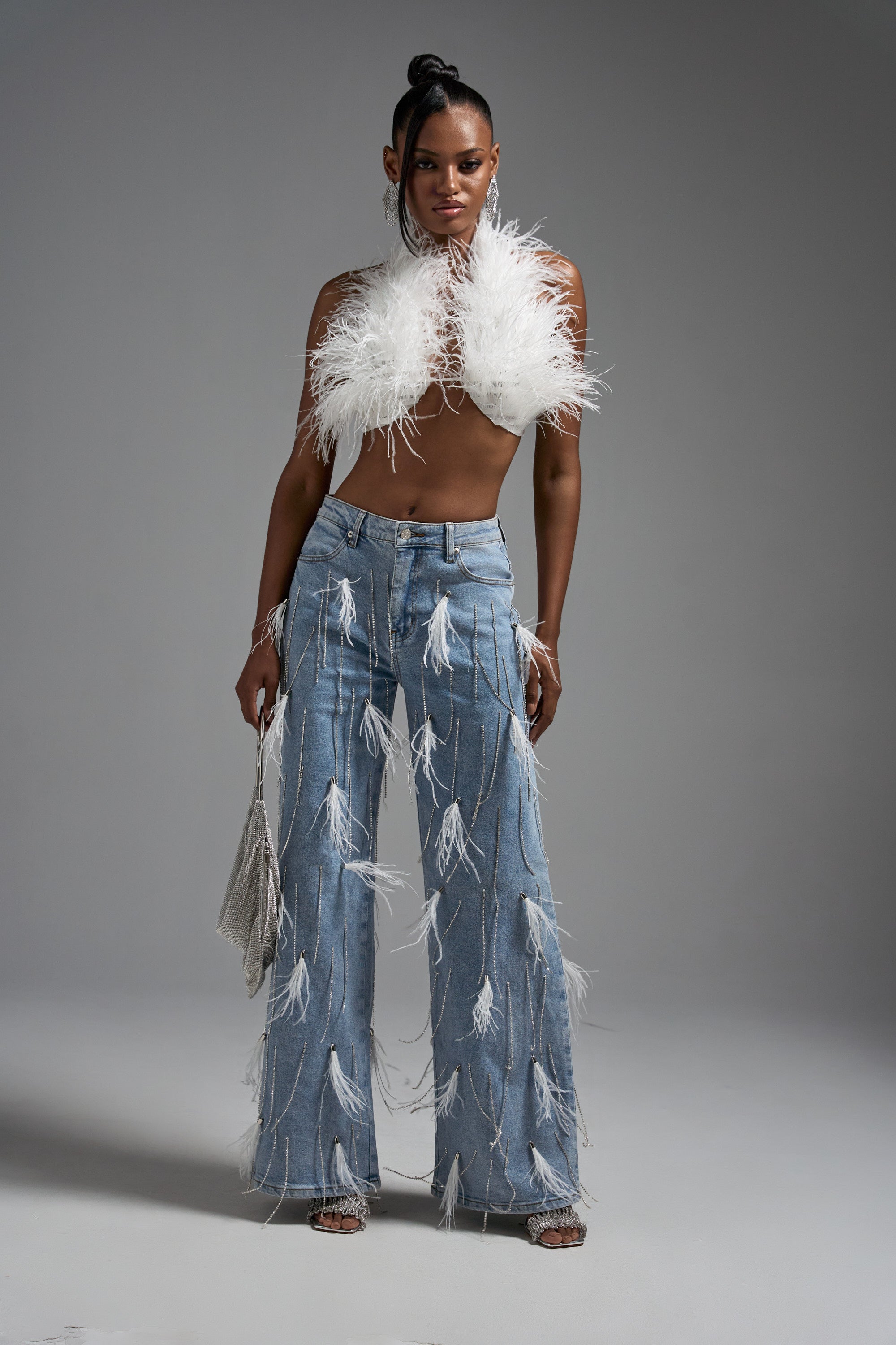 A woman stands confidently against a plain backdrop, wearing the SHAKE IT FOR ME GIRL FEATHER EMBELLISHED DENIM with a white feathered halter top, styled with a small bag and strappy heels.