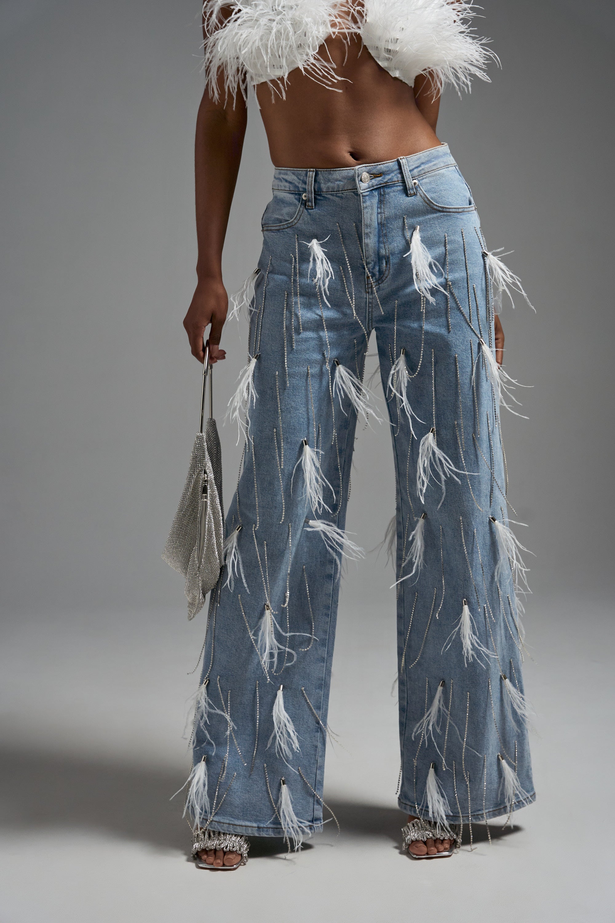 Wearing the SHAKE IT FOR ME GIRL FEATHER EMBELLISHED DENIM, which features white feather accents on high-rise relaxed fit jeans, paired with a matching feathered top and a silver handbag.