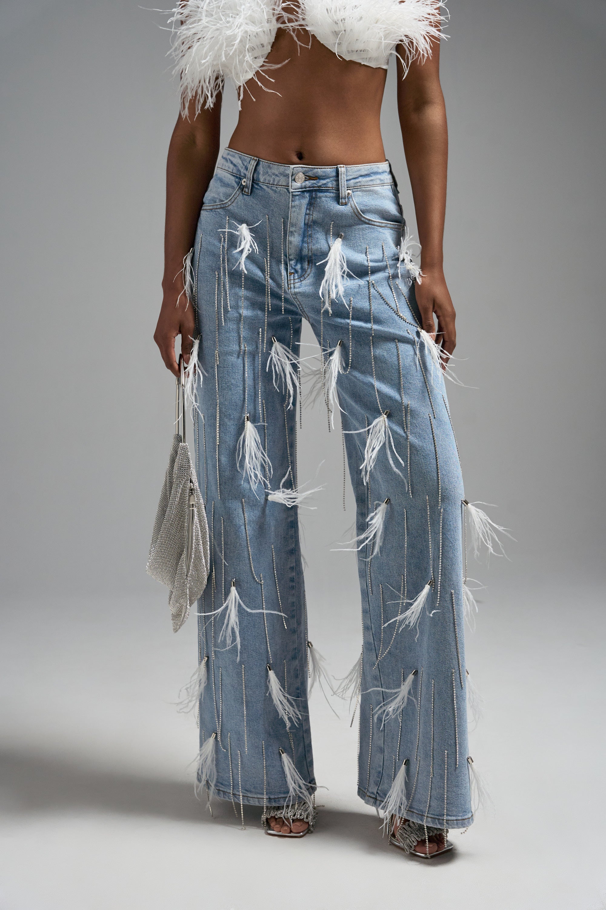 Wearing the SHAKE IT FOR ME GIRL FEATHER EMBELLISHED DENIM jeans, a white feathered crop top, and holding a small glittery bag, a person stands against a plain gray background.